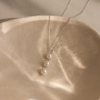 The Pearl Cascade Necklace in Sterling Silver, featuring three luminous pearls, rests on a textured, beige ceramic dish in soft light.