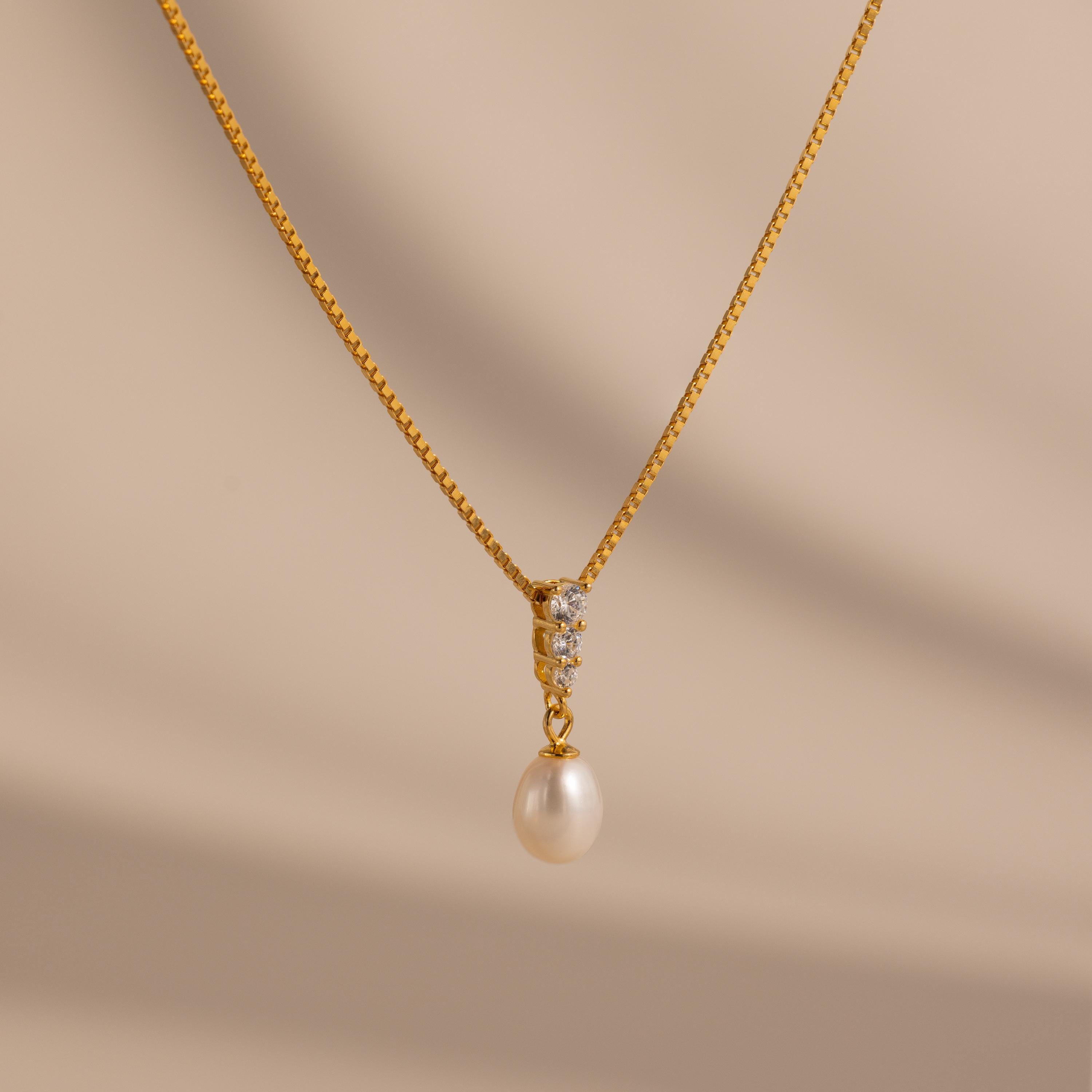The Diamond Pearl Droplet Necklace features a gold drop design with a pearl pendant and small diamonds, shown against a light beige background—an elegant jewelry piece suitable for any occasion.