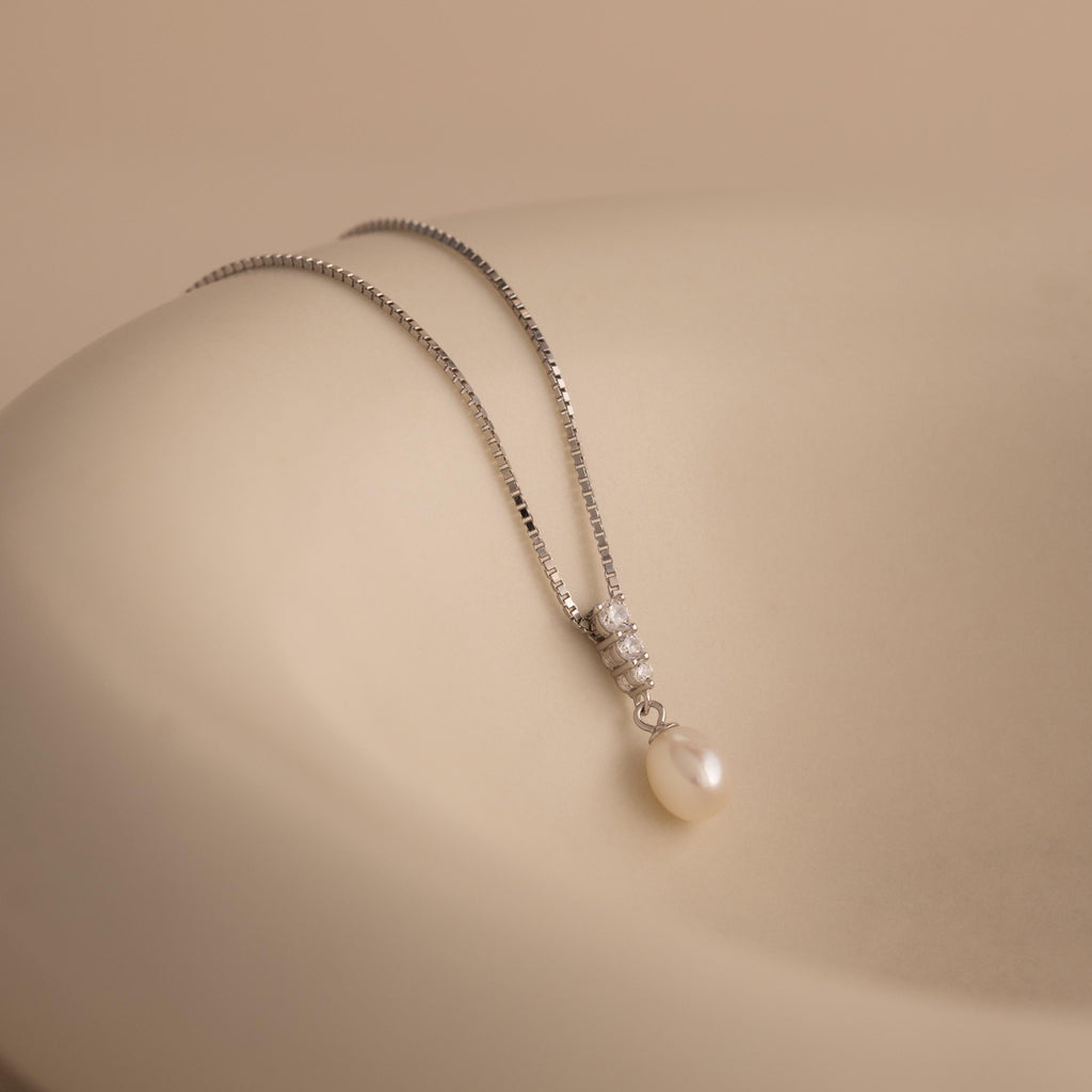 Diamond Pearl Droplet Necklace in Sterling Silver features a pearl drop pendant with small diamonds, displayed on a smooth, beige surface.