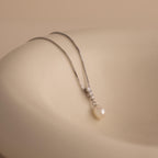 Diamond Pearl Droplet Necklace in Sterling Silver features a pearl drop pendant with small diamonds, displayed on a smooth, beige surface.