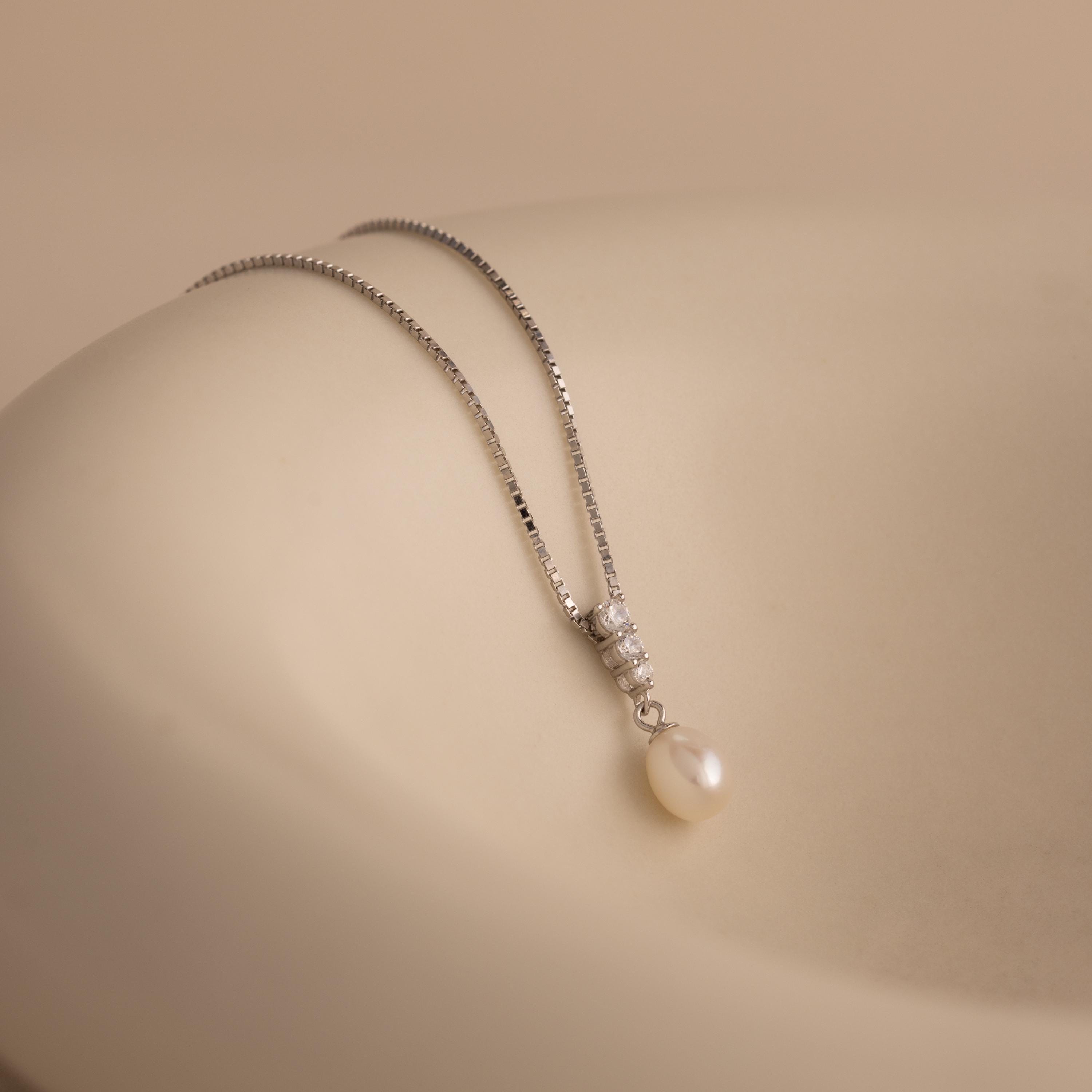 Diamond Pearl Droplet Necklace in Sterling Silver features a pearl drop pendant with small diamonds, displayed on a smooth, beige surface.