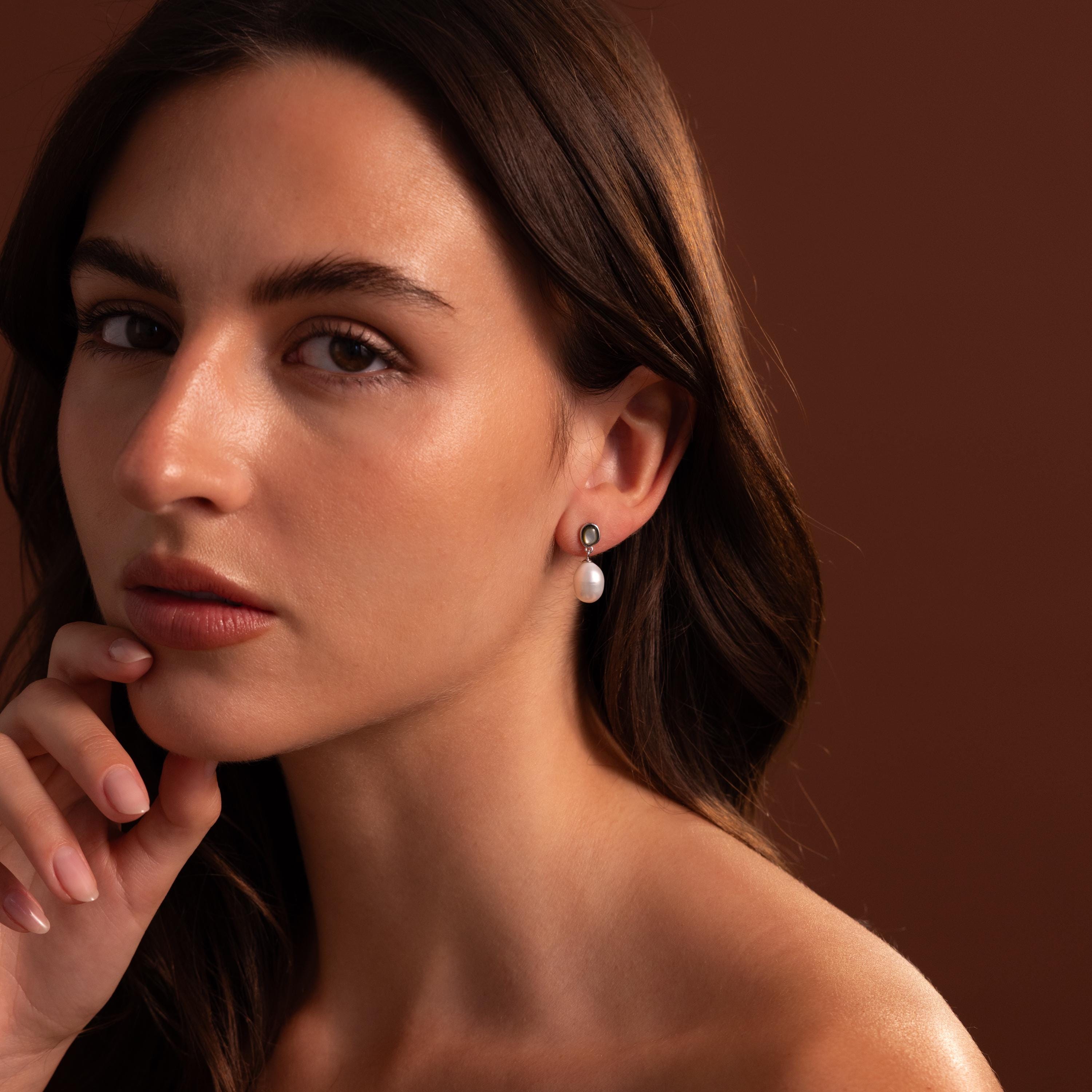 A woman with long brown hair gazes calmly, hand on chin, wearing Midnight Pearl Drop Earrings in Sterling Silver, against a soft brown background.