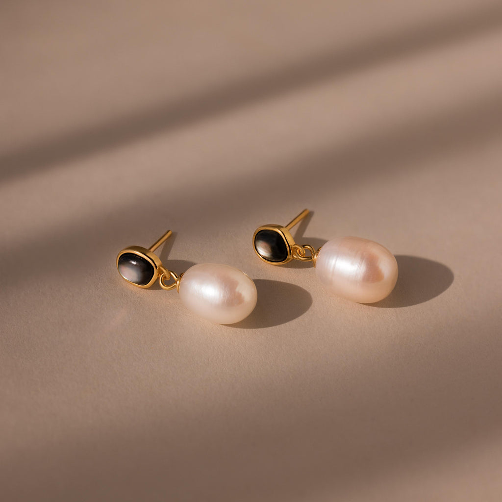 The Midnight Pearl Drop Earrings in 18K Gold showcase oval black accents and large white pearls for an elegant, timeless jewelry statement.