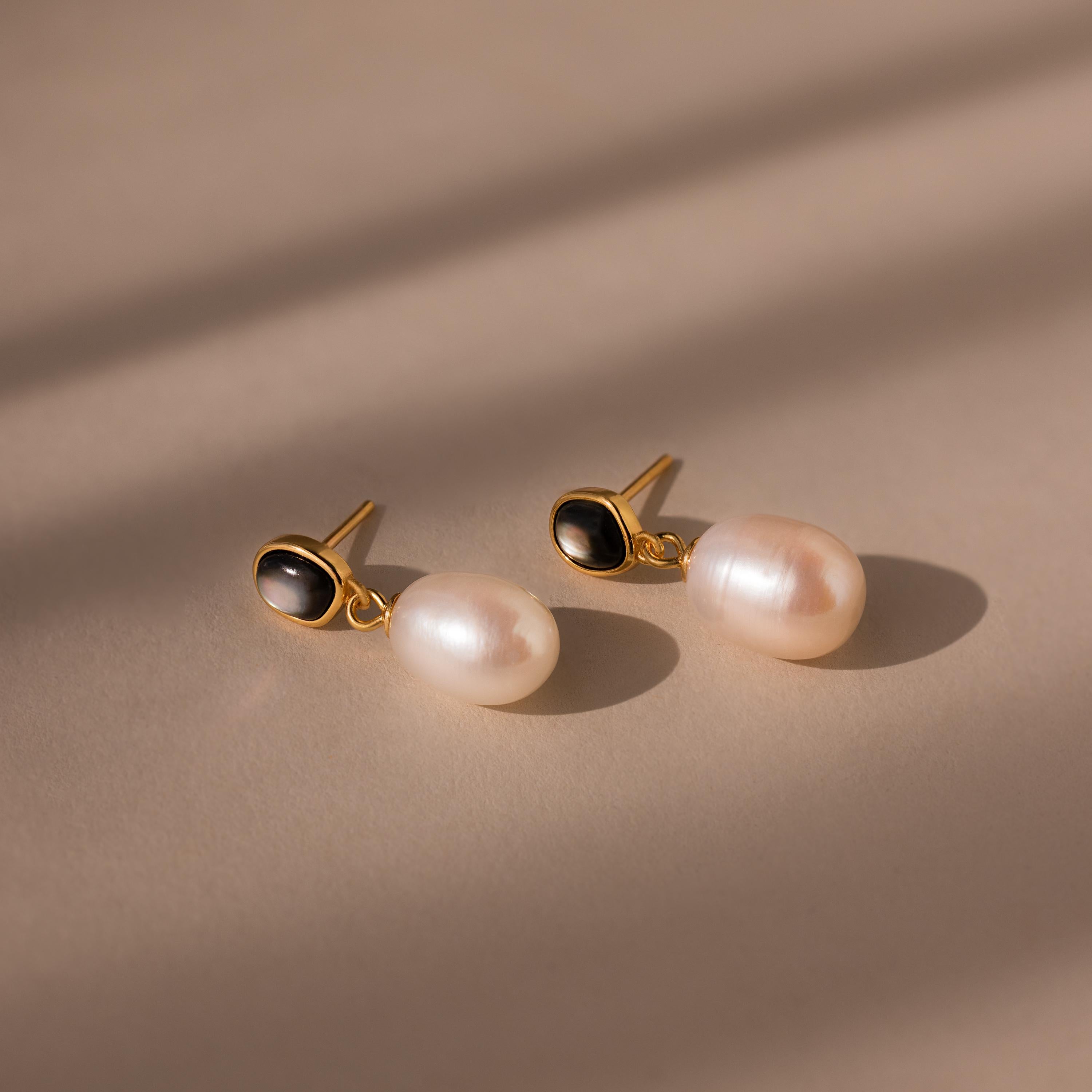 The Midnight Pearl Drop Earrings in 18K Gold showcase oval black accents and large white pearls for an elegant, timeless jewelry statement.