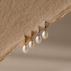 Two pairs of Diamond Pearl Droplet Earrings—one gold, one silver—with pearl drops and diamond-studded tops rest elegantly on beige fabric.