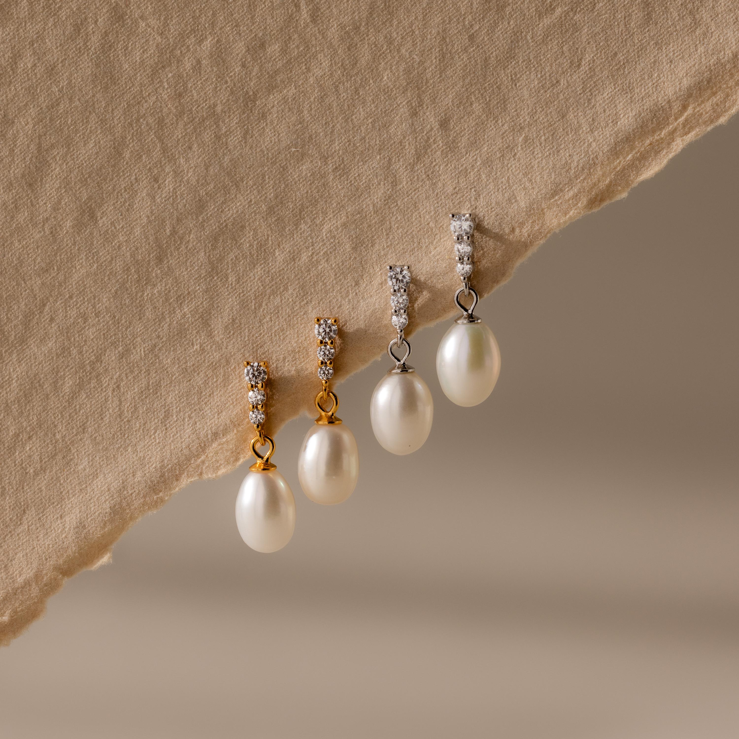 Two pairs of Diamond Pearl Droplet Earrings—one gold, one silver—with pearl drops and diamond-studded tops rest elegantly on beige fabric.