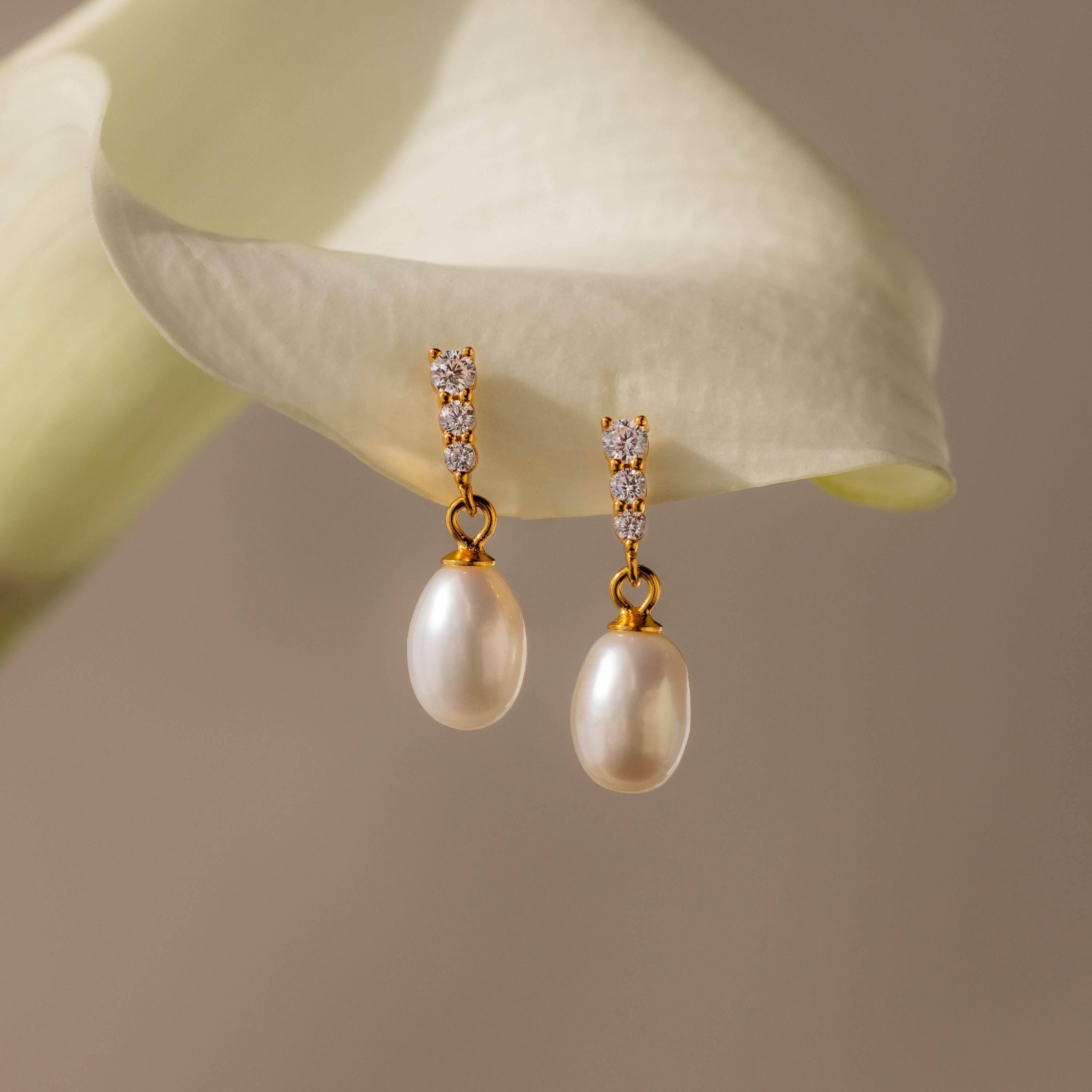 The Diamond Pearl Droplet Earrings feature gold and pearl drops with diamond accents, elegantly showcased against a white calla lily background.
