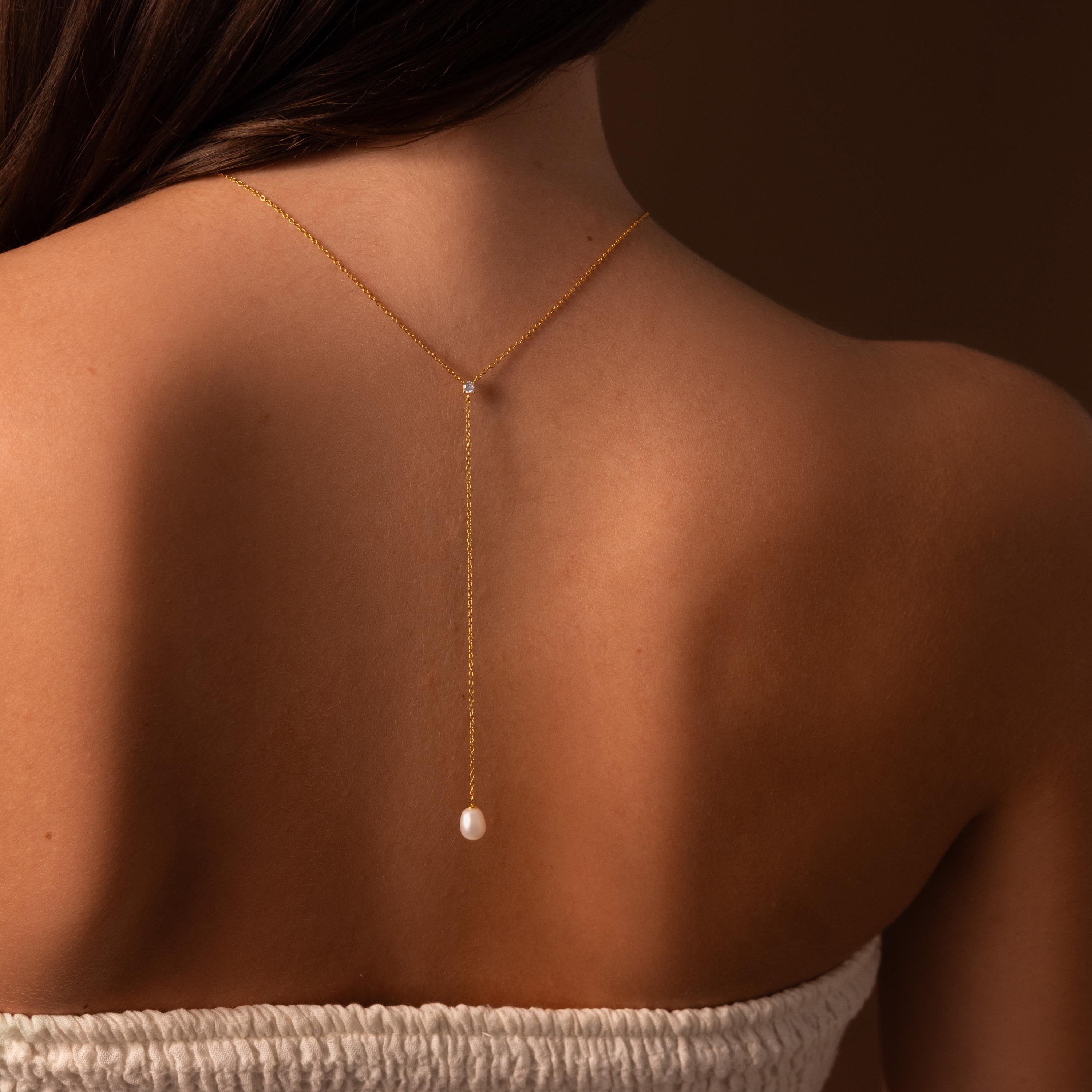 A woman wears the Pearl Drop Lariat Necklace, featuring a delicate vertical chain and a single pearl pendant that elegantly drapes down her bare back.