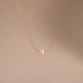 The Tiny Pearl Dot Necklace features a single white pearl pendant on a delicate gold chain, elegantly showcased against a soft beige background.