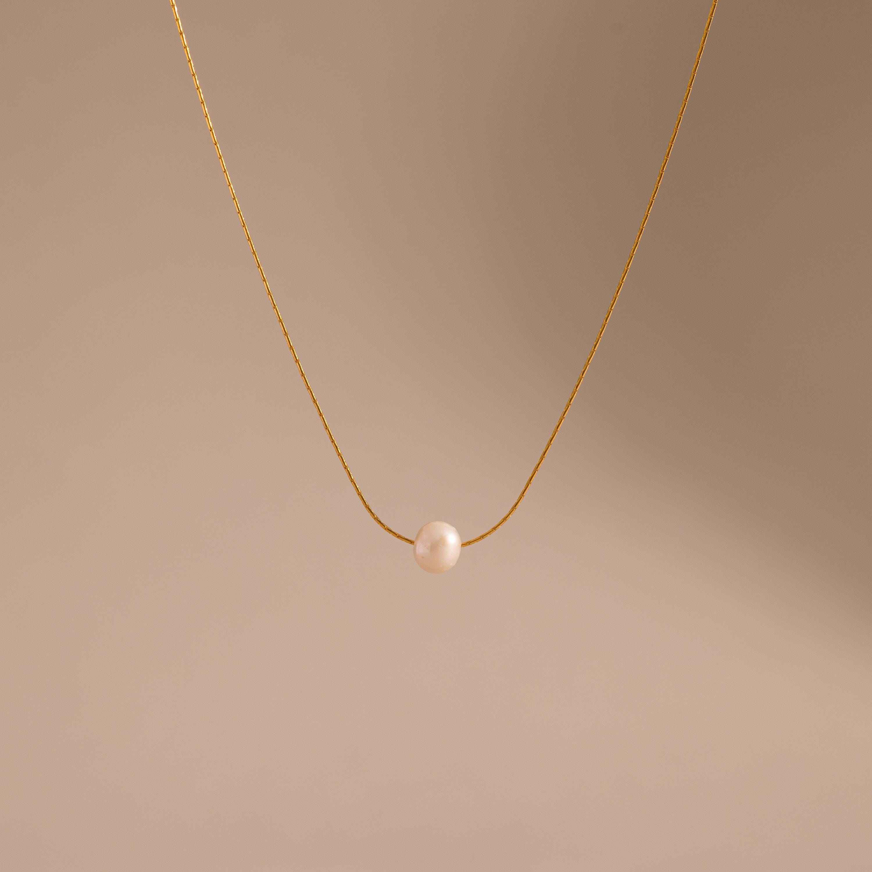The Tiny Pearl Dot Necklace features a single white pearl pendant on a delicate gold chain, elegantly showcased against a soft beige background.