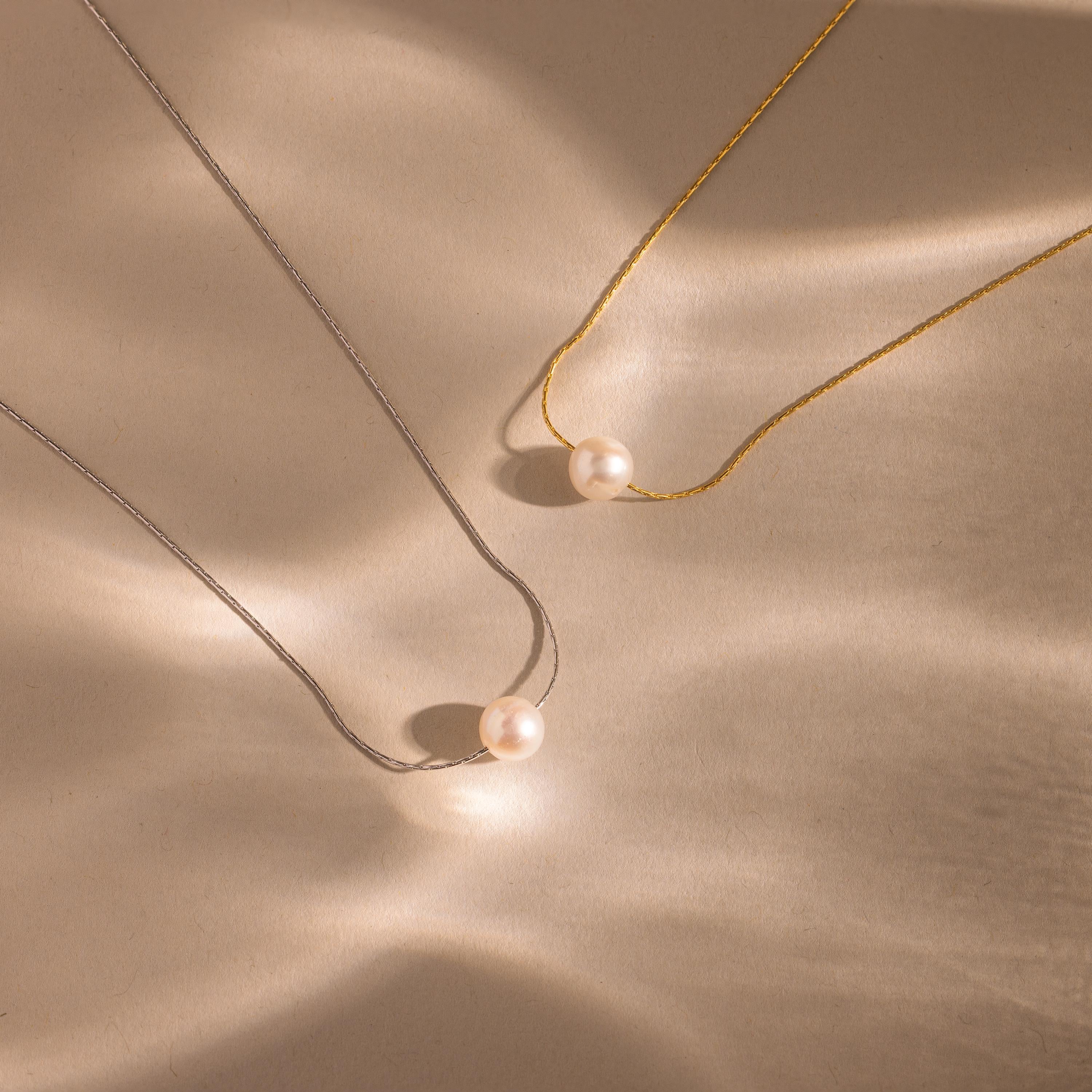 The Pearl Dot Necklace rests elegantly on a beige surface, its simple design highlighted by a delicate pearl accent.