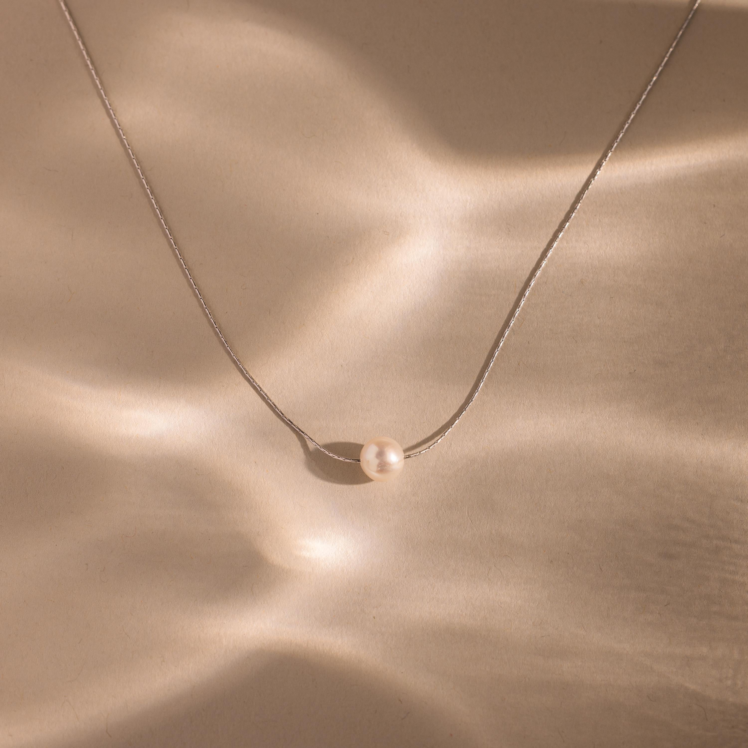 The Dainty Pearl Dot Necklace features a single pearl pendant on a delicate chain, displayed on a softly lit beige textured surface—ideal for layering with your favorite jewelry.