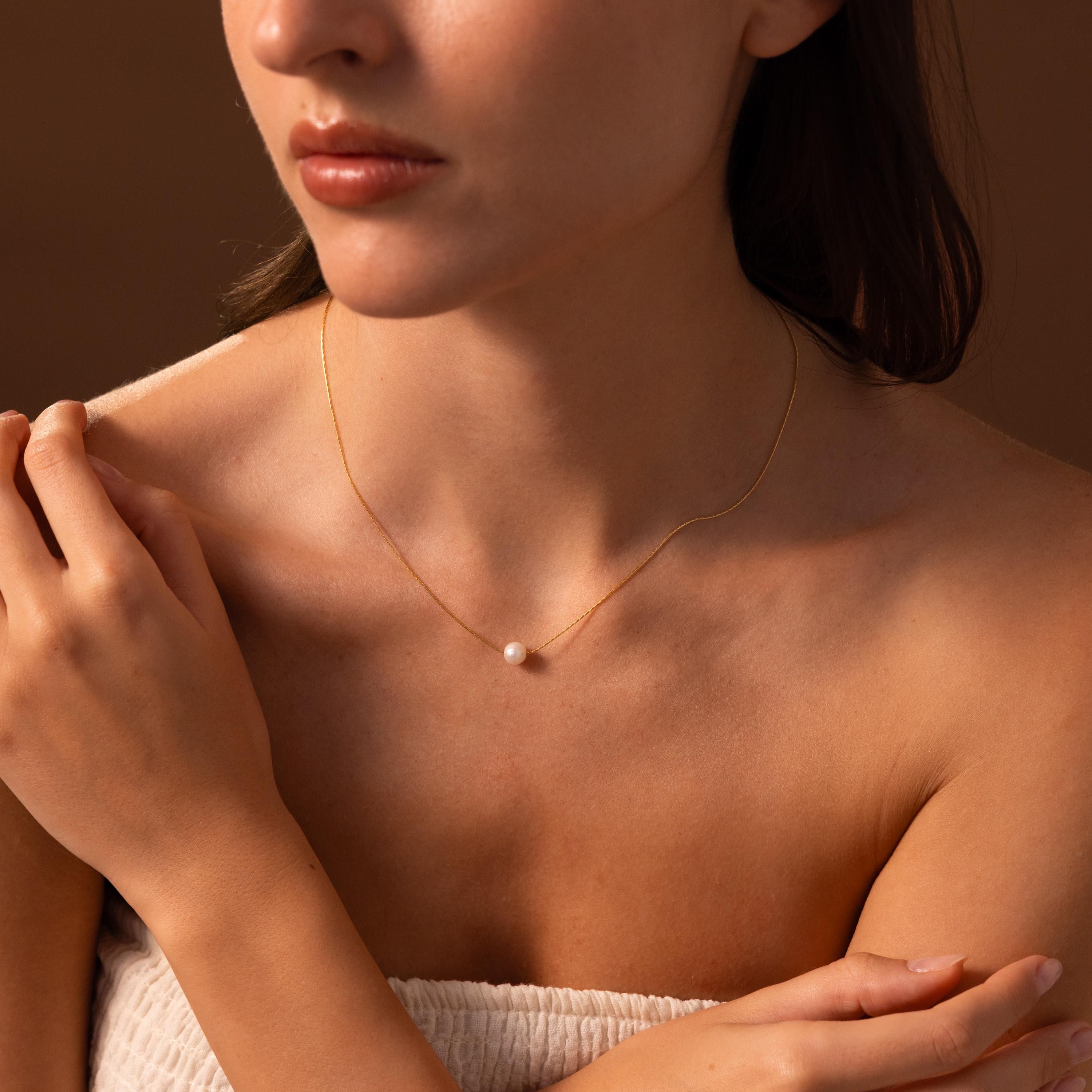 A woman wears the Dainty Pearl Dot Necklace—a simple gold chain with a single pearl—paired with a white textured top.