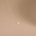 The Dainty Pearl Dot Necklace features a gold chain with a single round pearl pendant, shown on a beige background—ideal for layering or everyday wear.