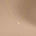 The Dainty Pearl Dot Necklace features a gold chain with a single round pearl pendant, shown on a beige background—ideal for layering or everyday wear.