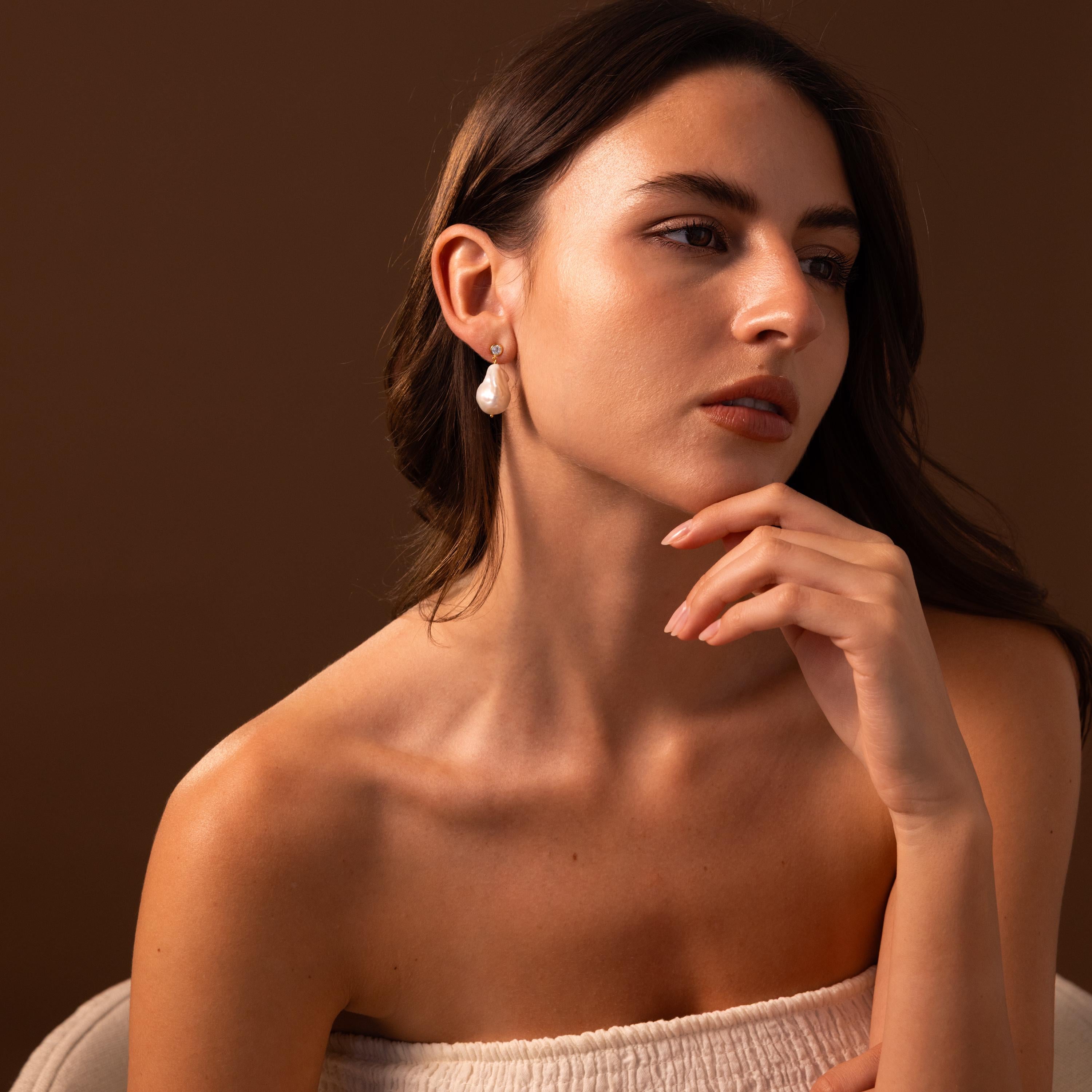 Woman with long brown hair wearing Diamond Baroque Pearl Studs and a white top, posing against a brown background.