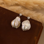 Diamond Baroque Pearl Studs with round crystal accents rest on a brown surface, pampas grass in the background—a stunning example of organic pearl jewelry.