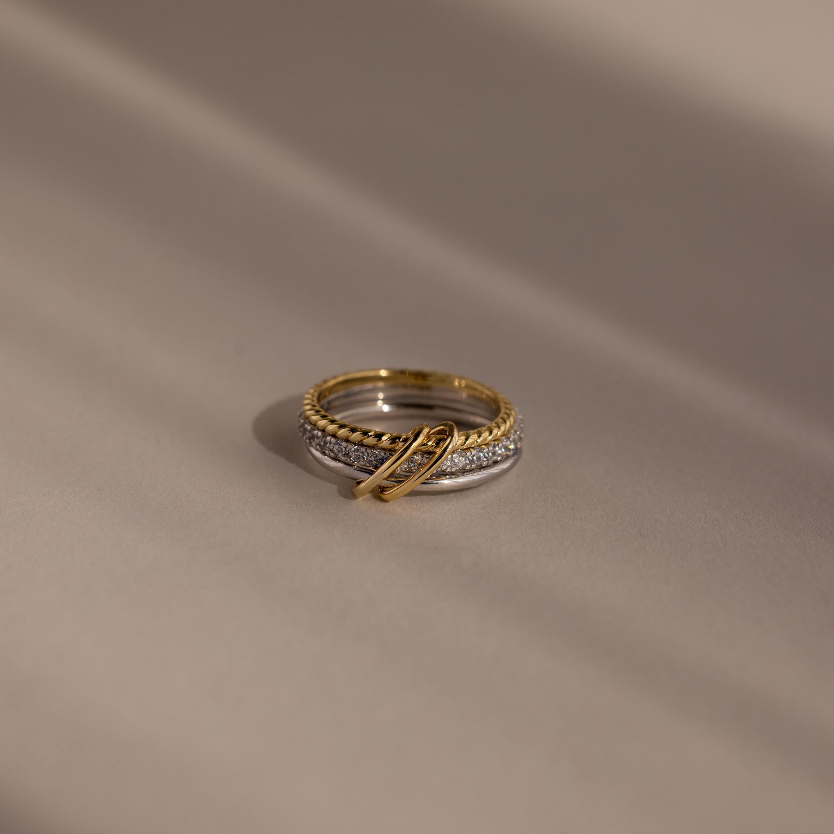 The Pave Mixed Metal Linked Ring Set, featuring gold and silver tones with diamond accents, rests on a smooth beige surface with soft shadows—a versatile piece for any occasion.