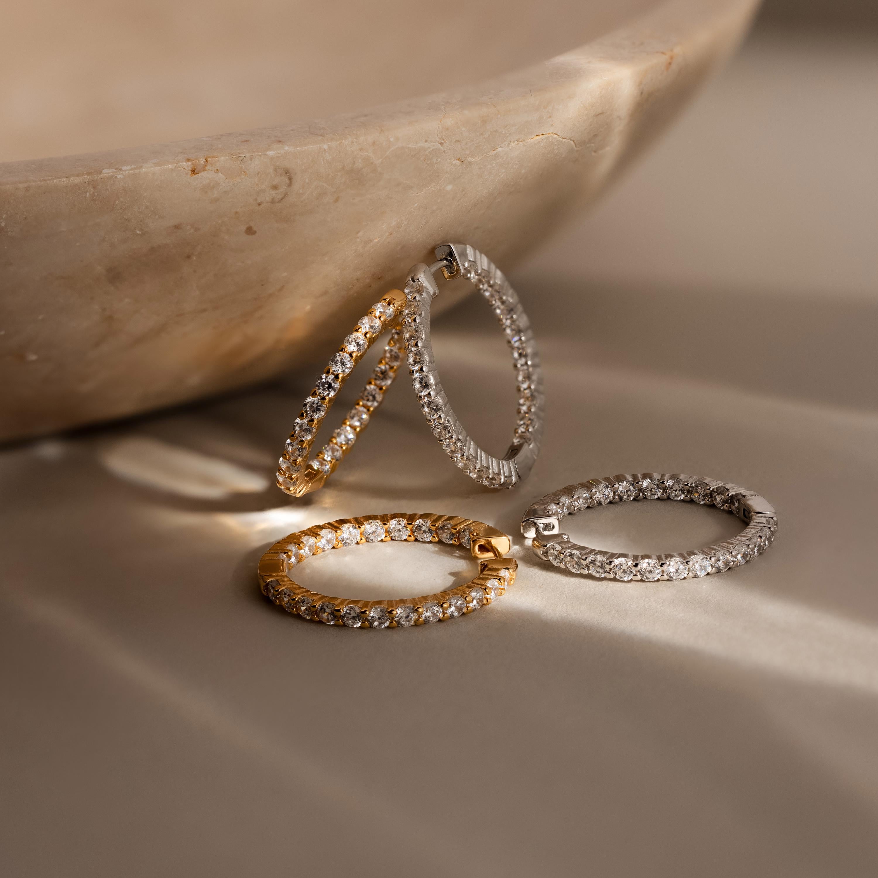 Four Inside Out Diamond Hoops—two in 18k gold and two in silver—are elegantly displayed on a beige stone surface.