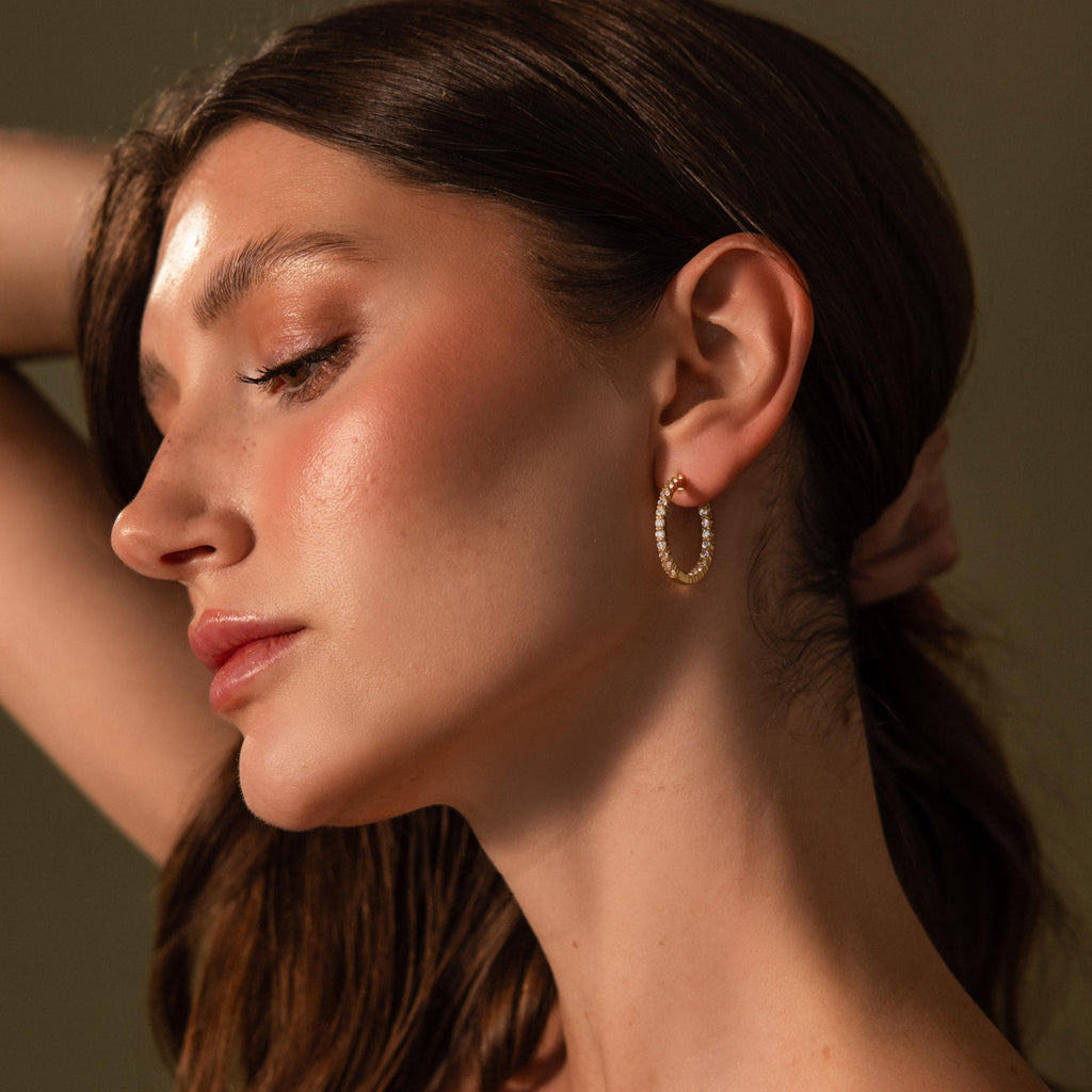 Woman with long brown hair wears sparkling Inside Out Diamond Hoops in 18k gold, posing with her arm raised behind her head.