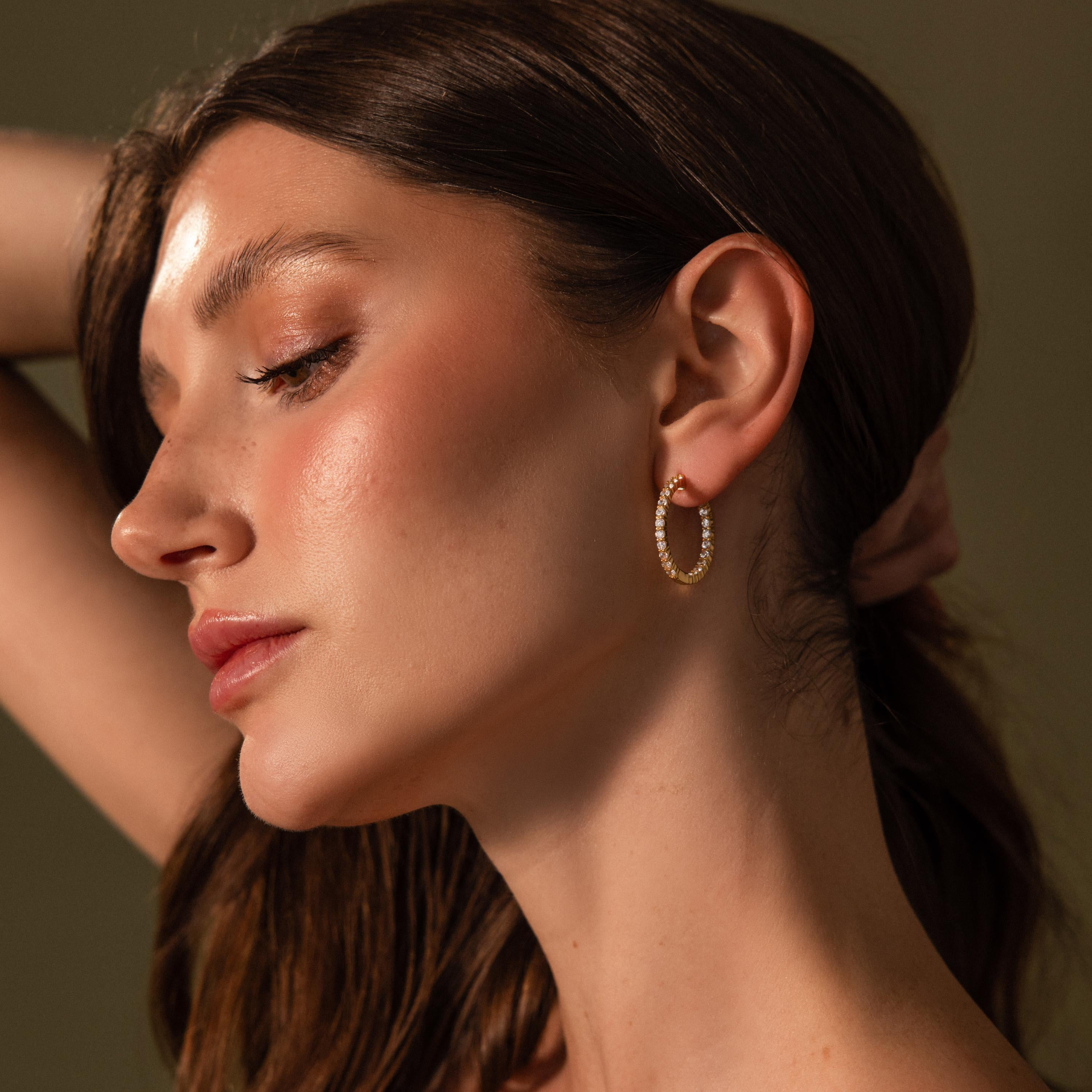 Woman with long brown hair wears sparkling Inside Out Diamond Hoops in 18k gold, posing with her arm raised behind her head.