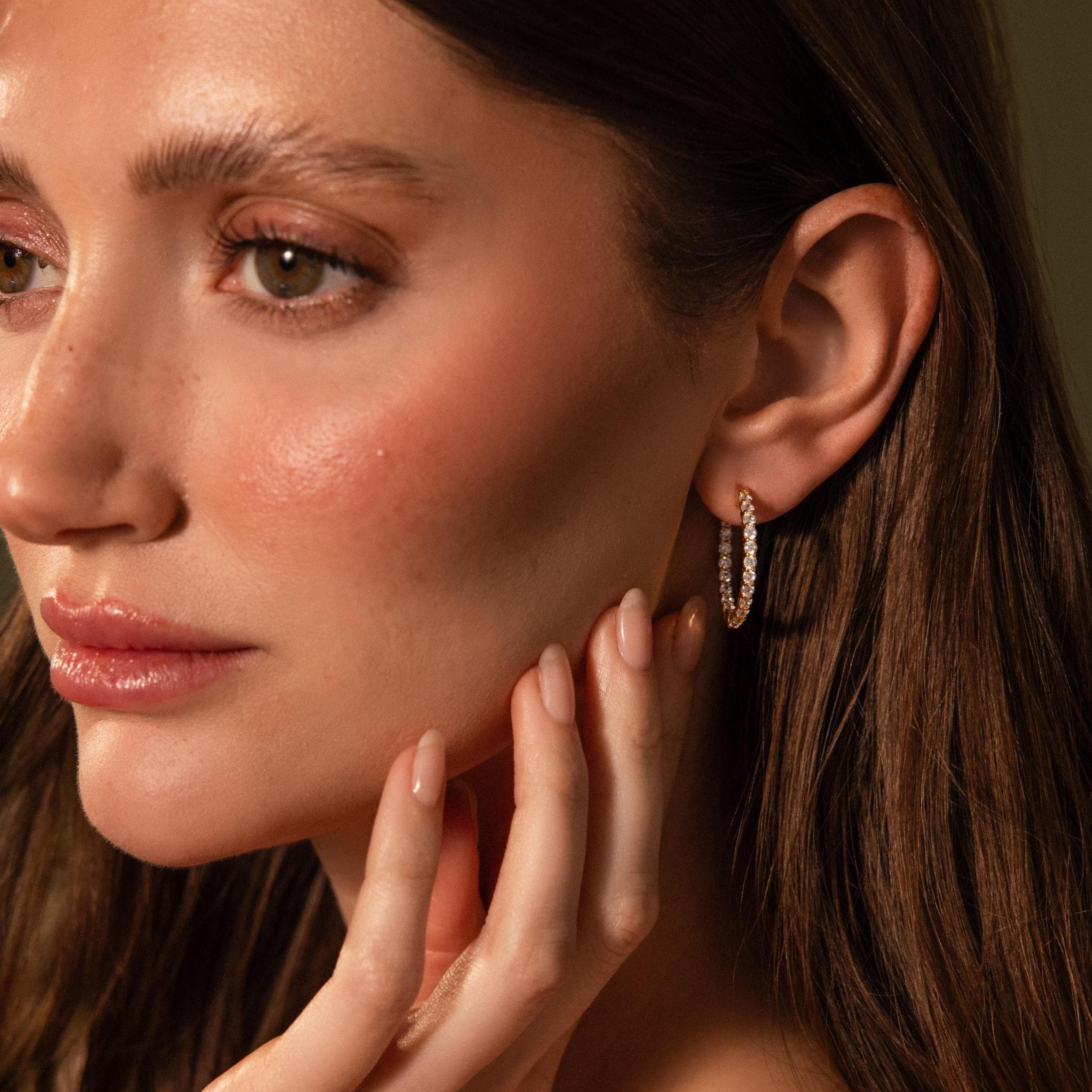 A woman with long brown hair wears Inside Out Diamond Hoops in 18K Gold, touching her face with manicured fingers.