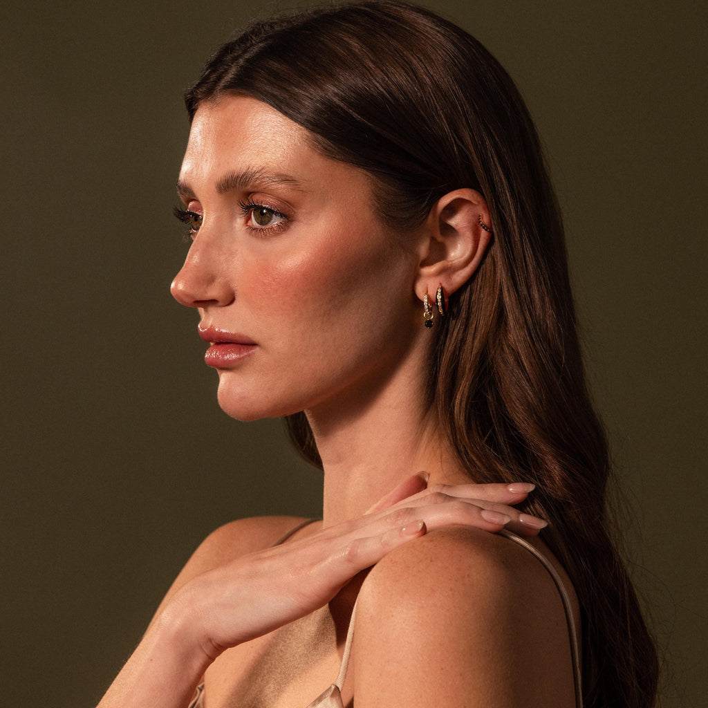 A woman with long brown hair models the Noir Pave Dome Hoops—gold earrings featuring black enamel accents and pave details—as she poses in profile against a dark background.