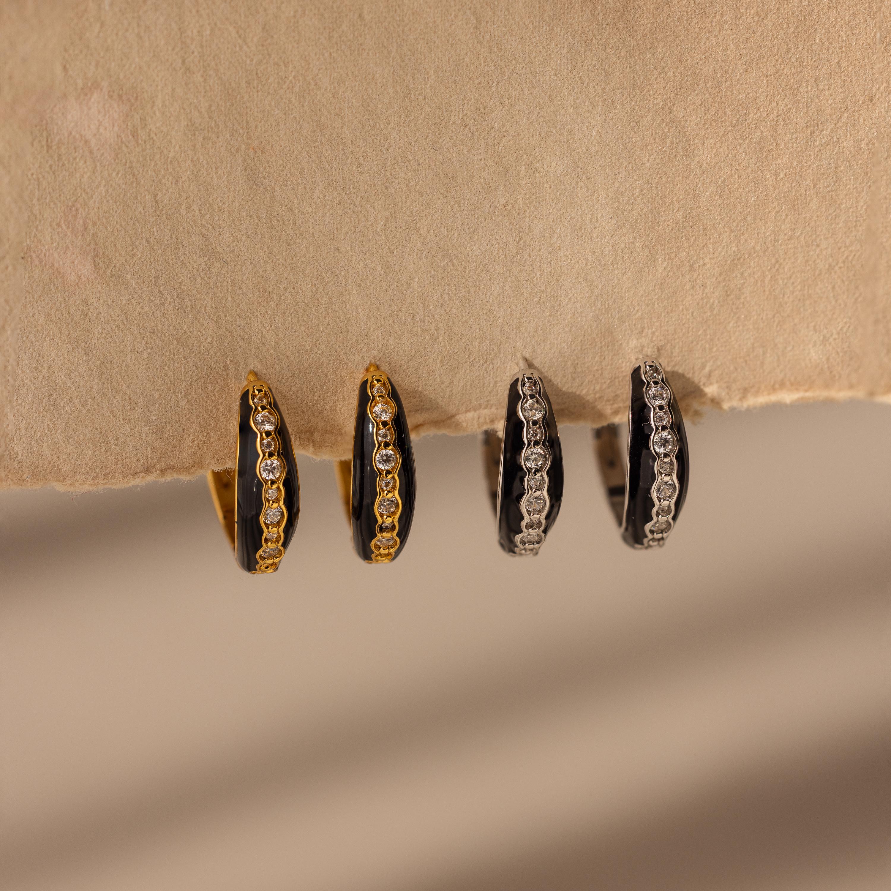 Four Noir Pave Dome Hoops earrings—two gold, two silver—with embedded gemstones and pave detailing are displayed on a beige textured surface.