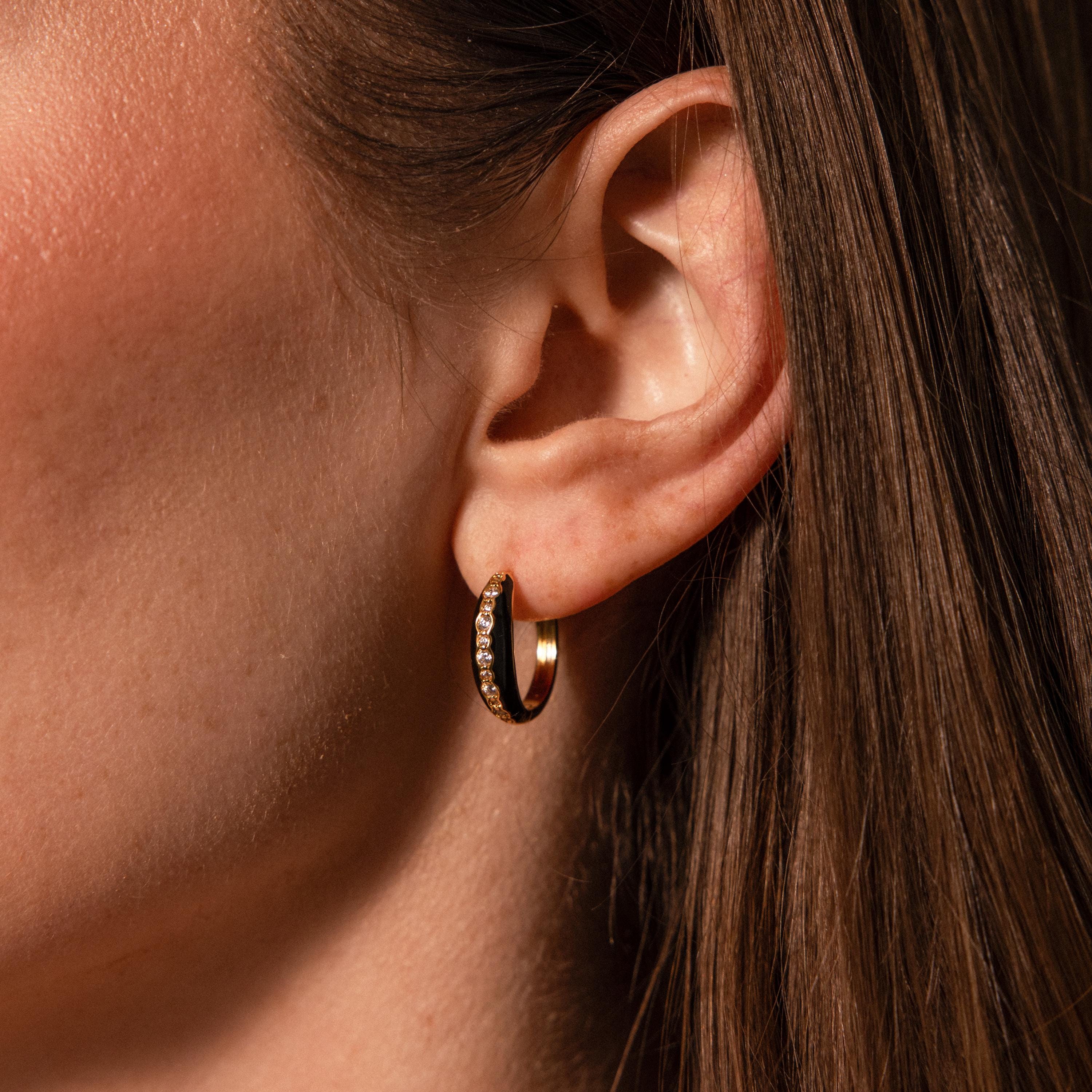 Close-up of a woman's ear wearing Noir Pave Dome Hoops in 18K Gold—glamorous gold hoops with sparkling stones and a sleek black enamel accent.