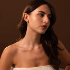 A woman with long brown hair, wearing a strapless white top, looks to the side against a brown background. She wears Pearl Bezel Studs in 18K Gold, adding an elegant touch perfect for everyday wear.