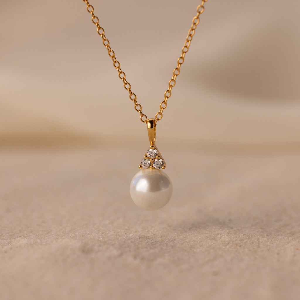 The Diamond Halo Pearl Necklace features a pearl pendant surrounded by small diamonds, elegantly showcased on a sandy surface—ideal for those who value timeless jewelry.