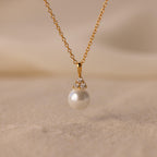 The Diamond Halo Pearl Necklace features a pearl pendant surrounded by small diamonds, elegantly showcased on a sandy surface—ideal for those who value timeless jewelry.