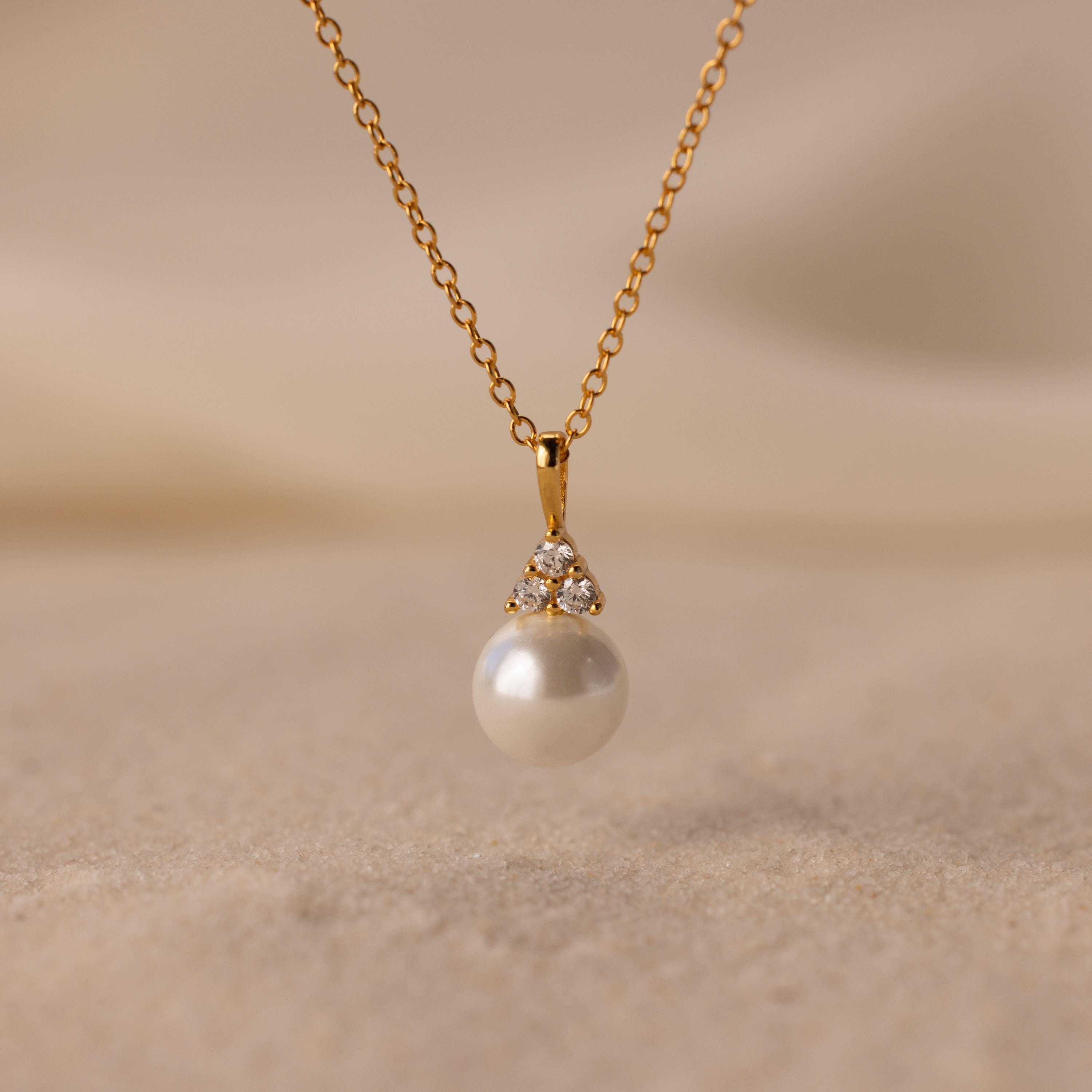 The Diamond Halo Pearl Necklace features a pearl pendant surrounded by small diamonds, elegantly showcased on a sandy surface—ideal for those who value timeless jewelry.