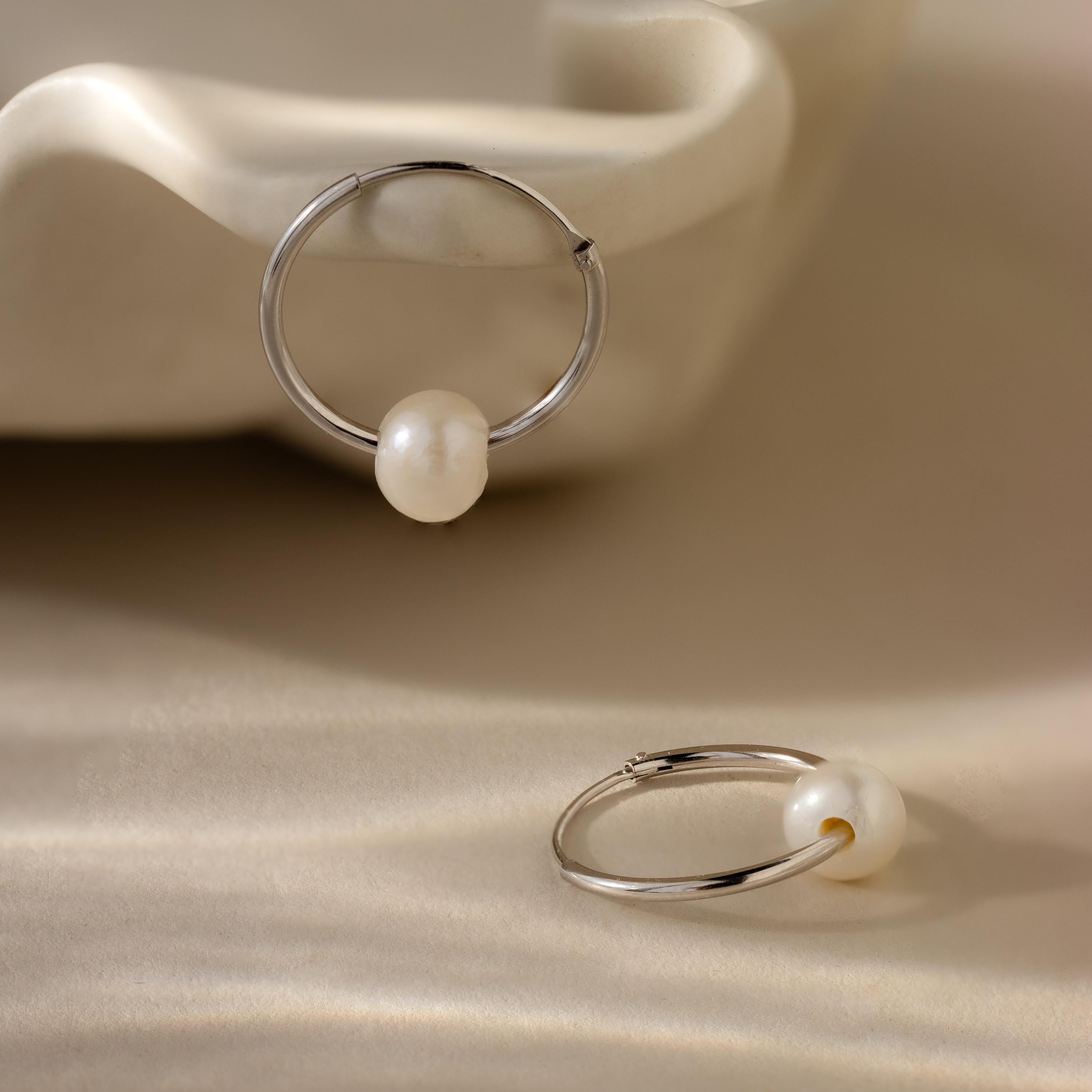 Two Round Pearl Hoops in Sterling Silver, each featuring a luminous pearl, are displayed on a soft beige surface.