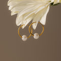 The Round Pearl Hoops feature gold hoops with pearls hanging from a white flower, set against a neutral background—an elegant, timeless jewelry choice.