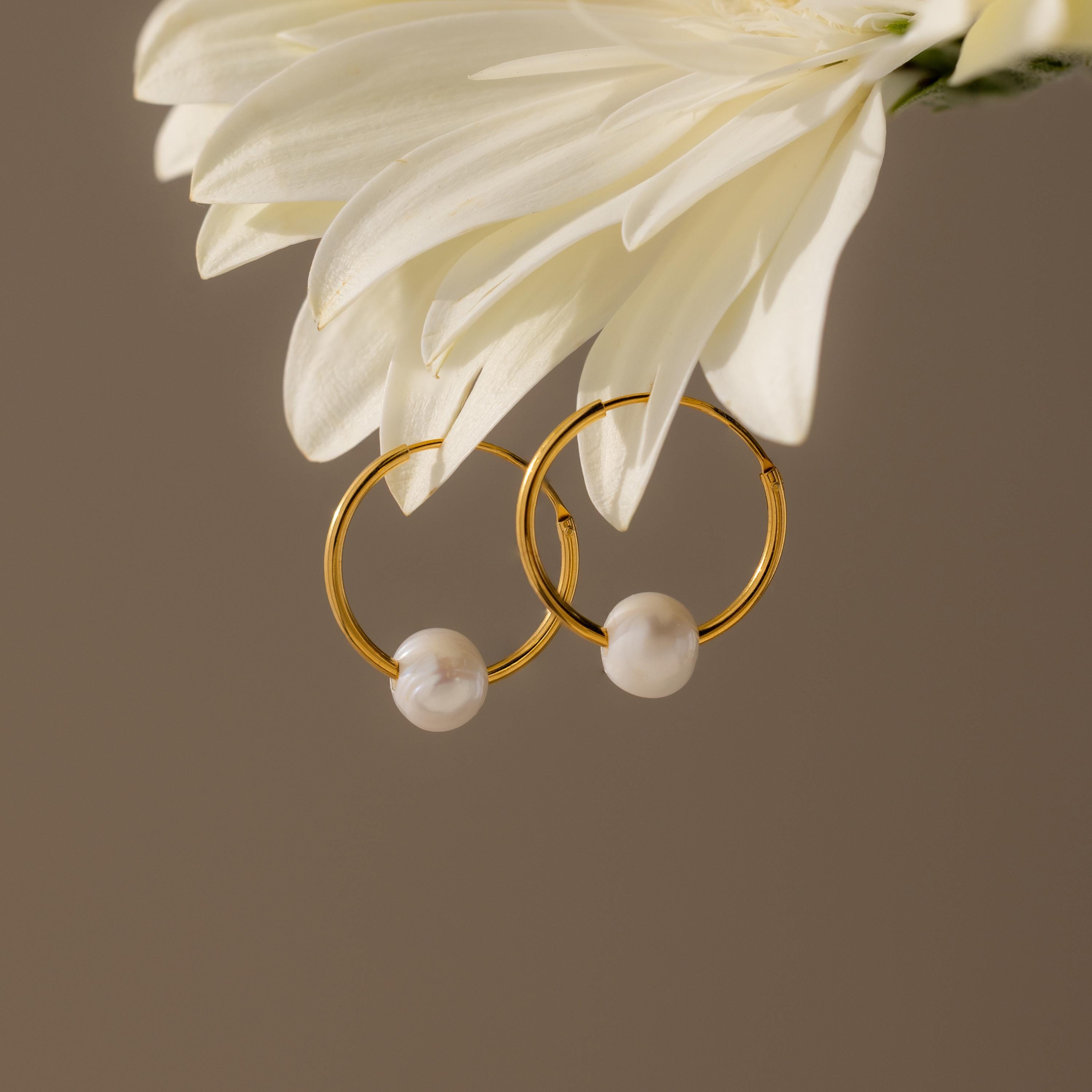 The Round Pearl Hoops feature gold hoops with pearls hanging from a white flower, set against a neutral background—an elegant, timeless jewelry choice.