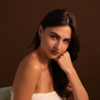 Woman with long dark hair in a white top rests her chin on her hand against a brown background, accessorized with minimal jewelry featuring elegant Round Pearl Hoops in 18K Gold.