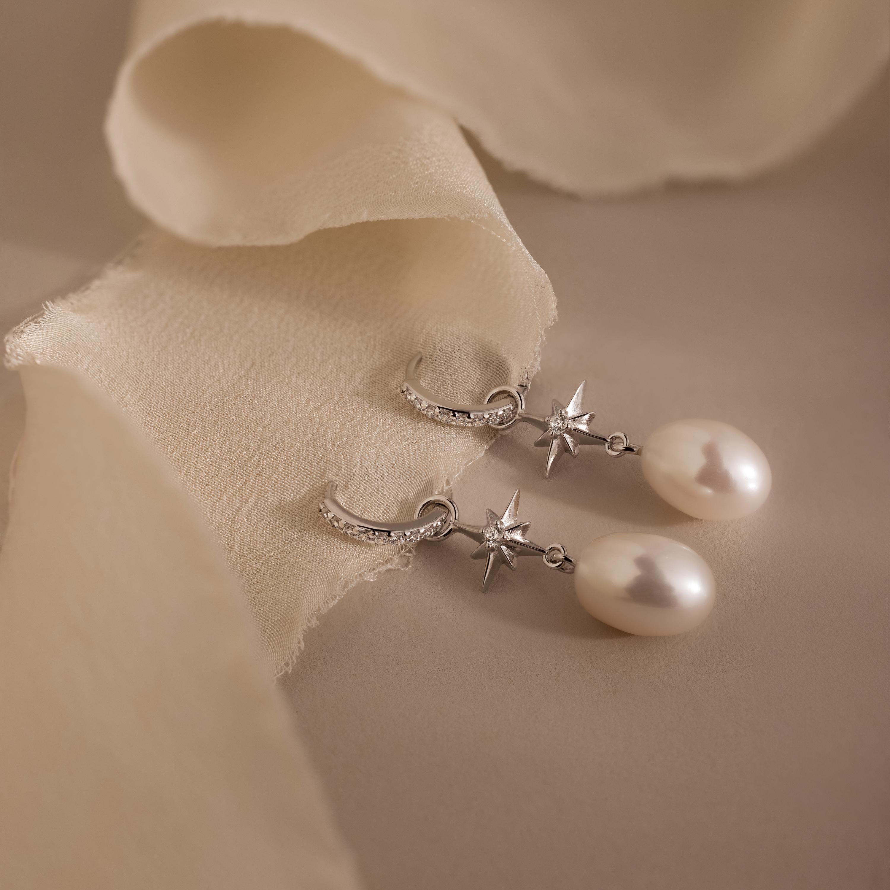 North Star Pearl Hoops adorned with star accents and delicate pearls, showcased on a soft beige fabric background.