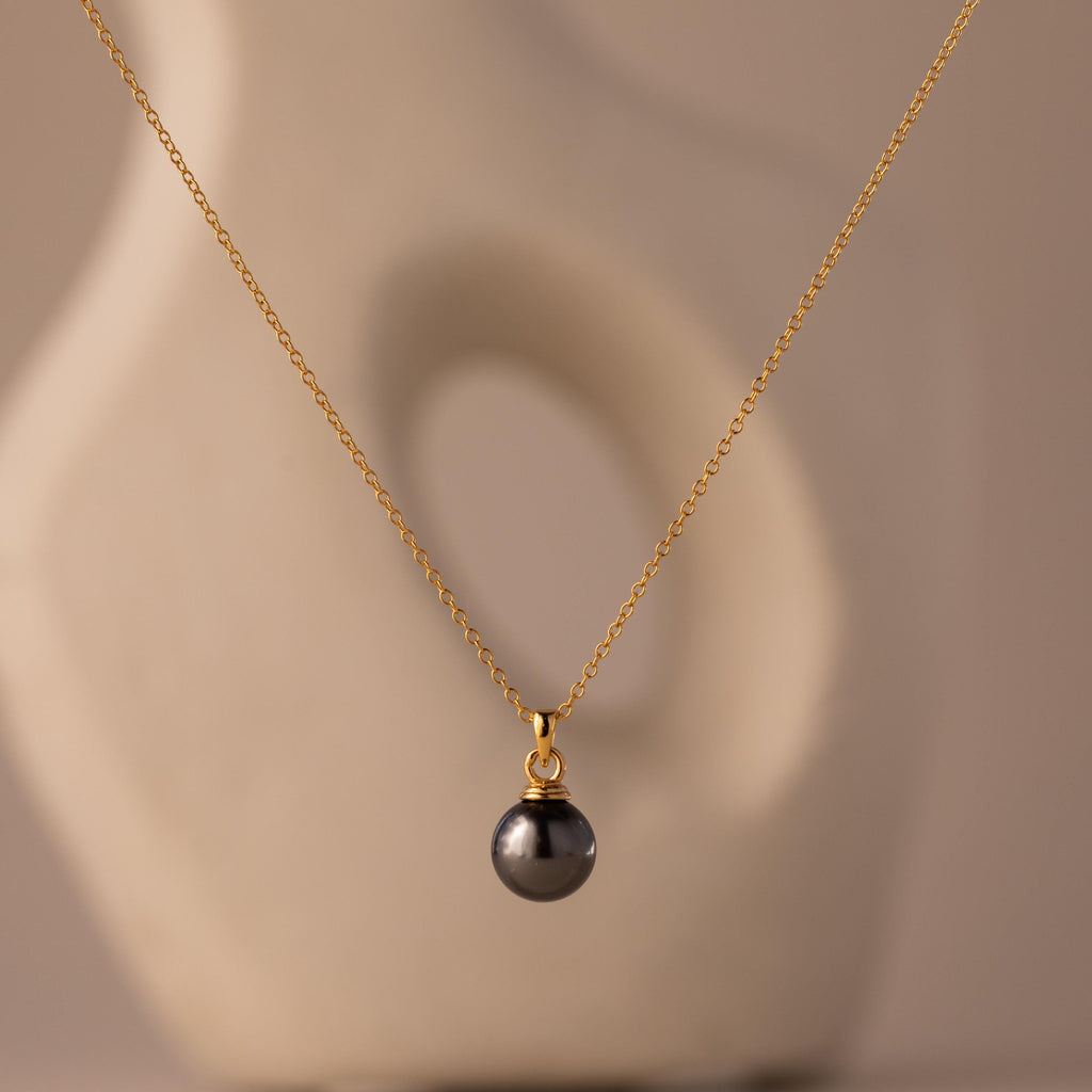 The Midnight Pearl Necklace features a gold chain with a single dark pearl pendant, captured against a soft, abstract beige background.
