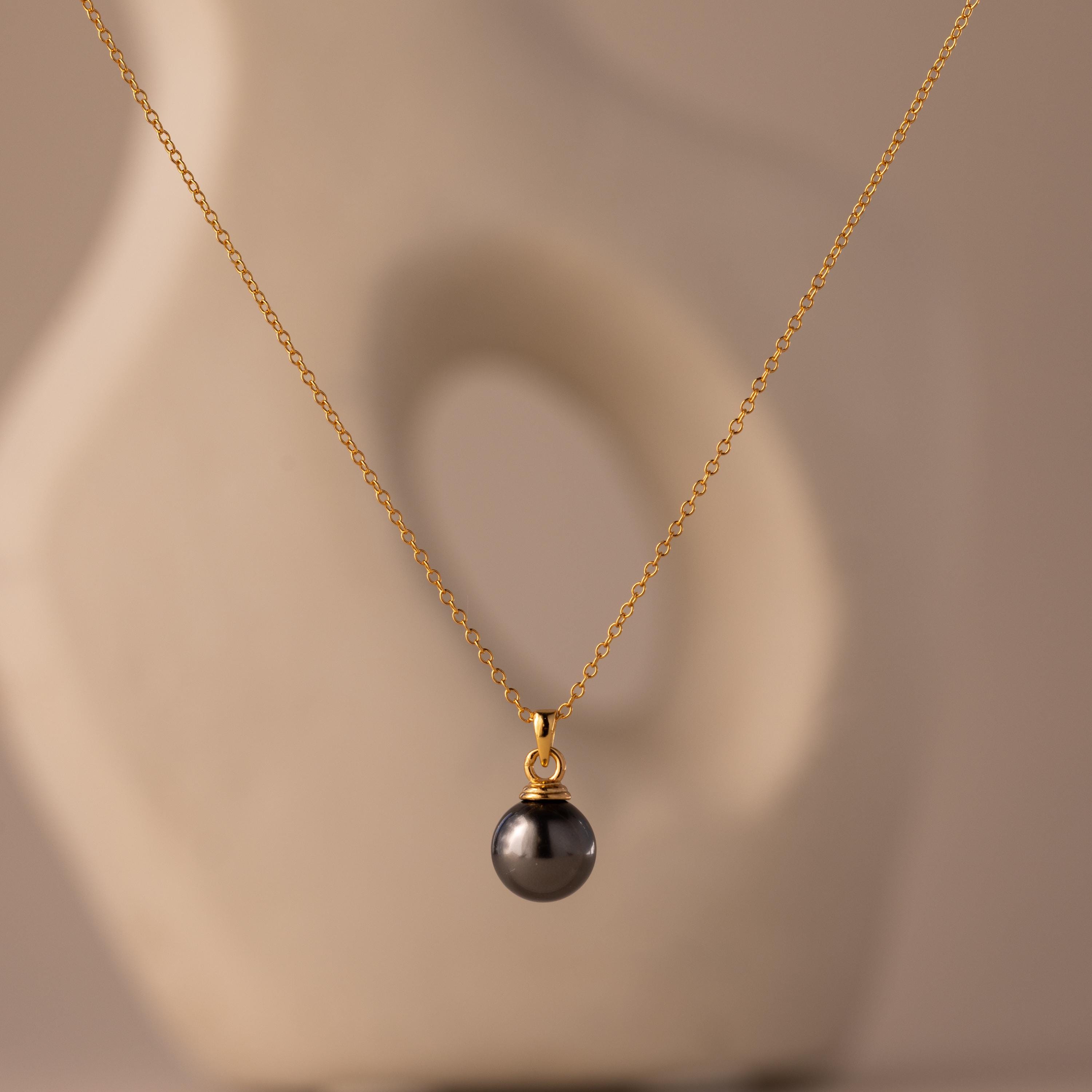 The Midnight Pearl Necklace features a gold chain with a single dark pearl pendant, captured against a soft, abstract beige background.