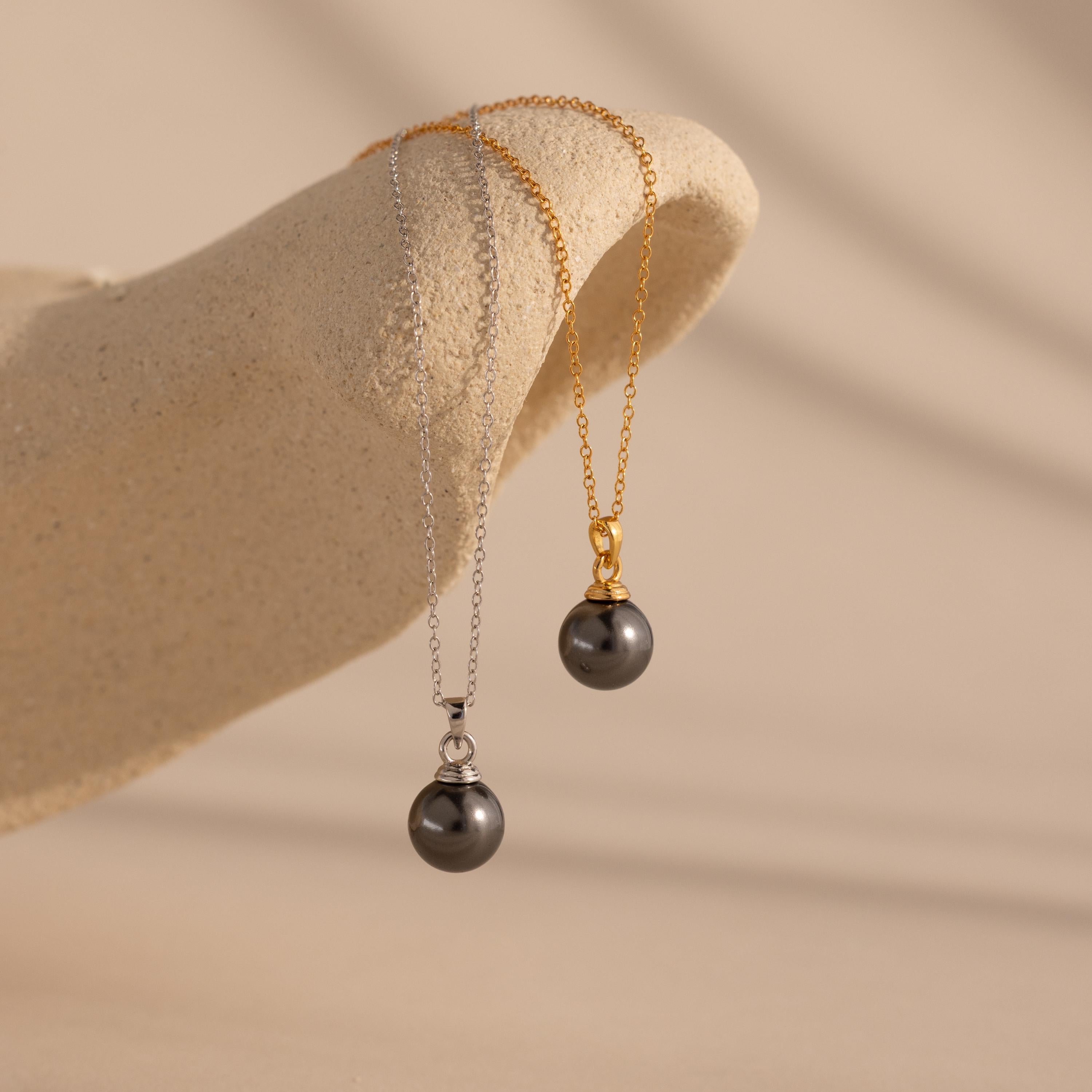 The Midnight Pearl Necklace, offered with silver and gold chains, is elegantly displayed on a textured beige surface—timeless jewelry suited for any occasion.
