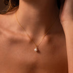 A close-up of a woman's neck showcases the Diamond Halo Pearl Necklace—a gold chain with a single pearl pendant, embodying timeless elegance.