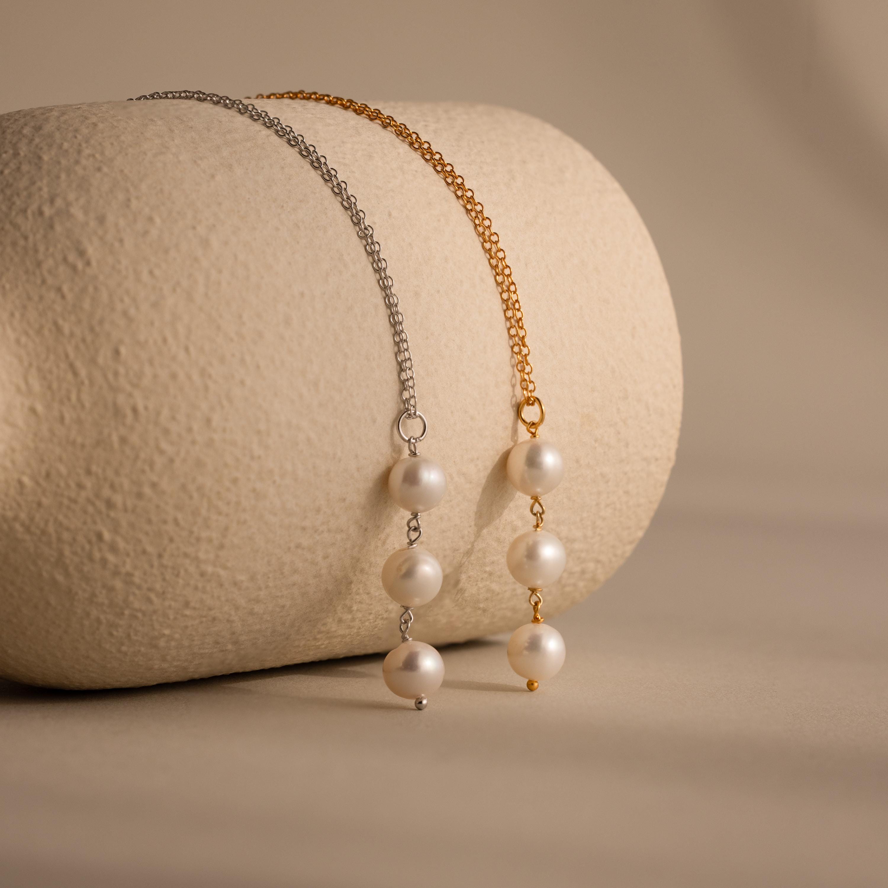 Two Pearl Cascade Necklaces, one with a silver chain and one with gold, are elegantly displayed on a textured beige roll.