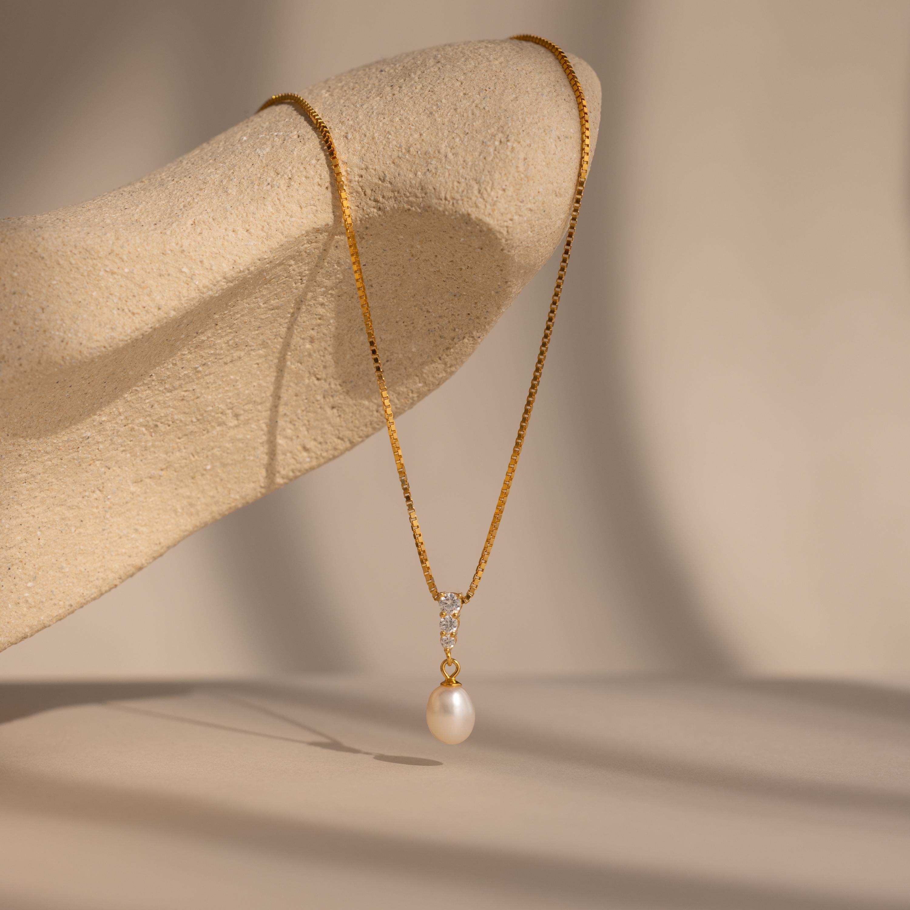 The Diamond Pearl Droplet Necklace in 18K Gold is elegantly draped over a textured beige sculpture, showcasing its gold chain and pearl pendant.