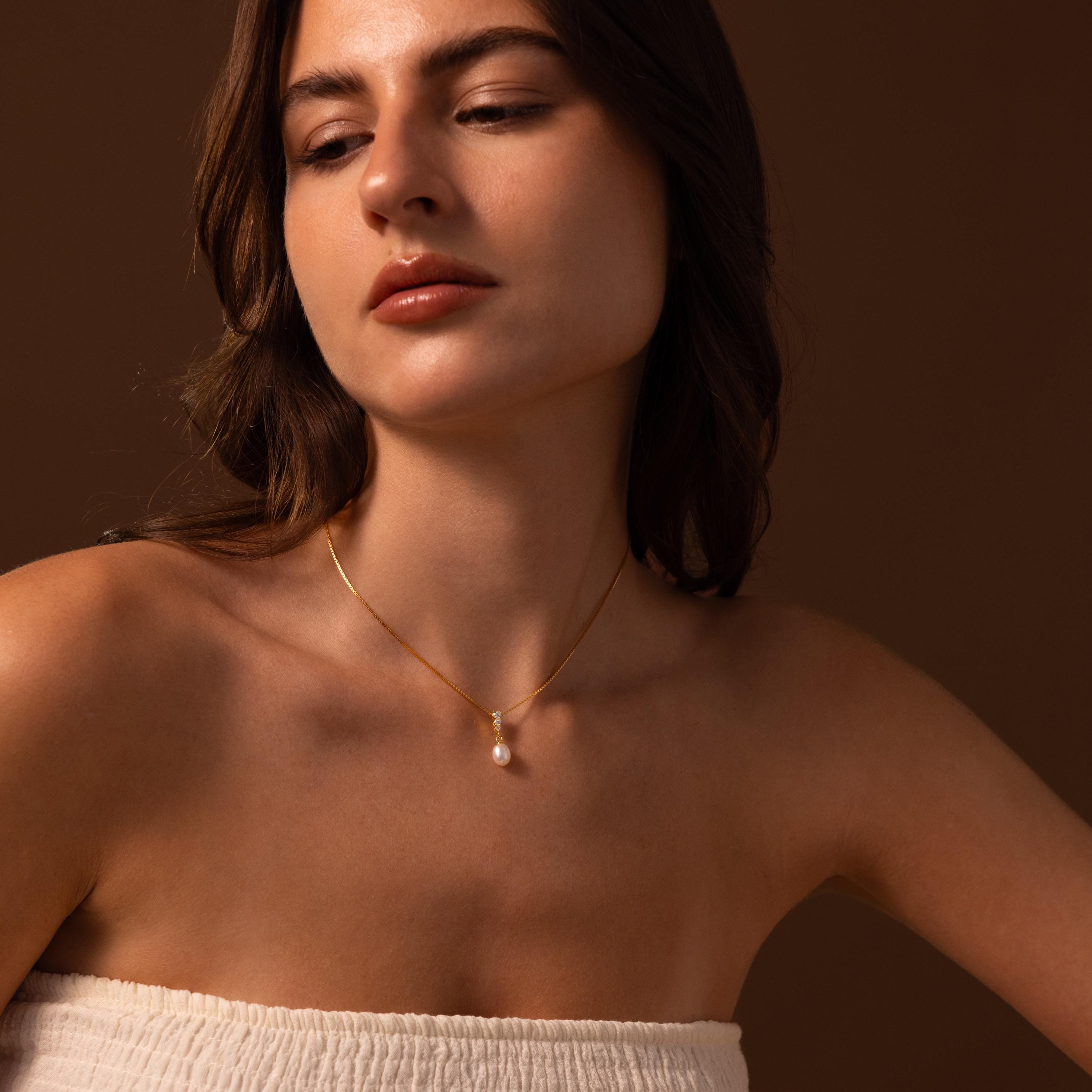 Woman with wavy brown hair wearing the Diamond Pearl Droplet Necklace in 18K Gold, featuring a pearl pendant, and a white strapless top, set against a brown background.