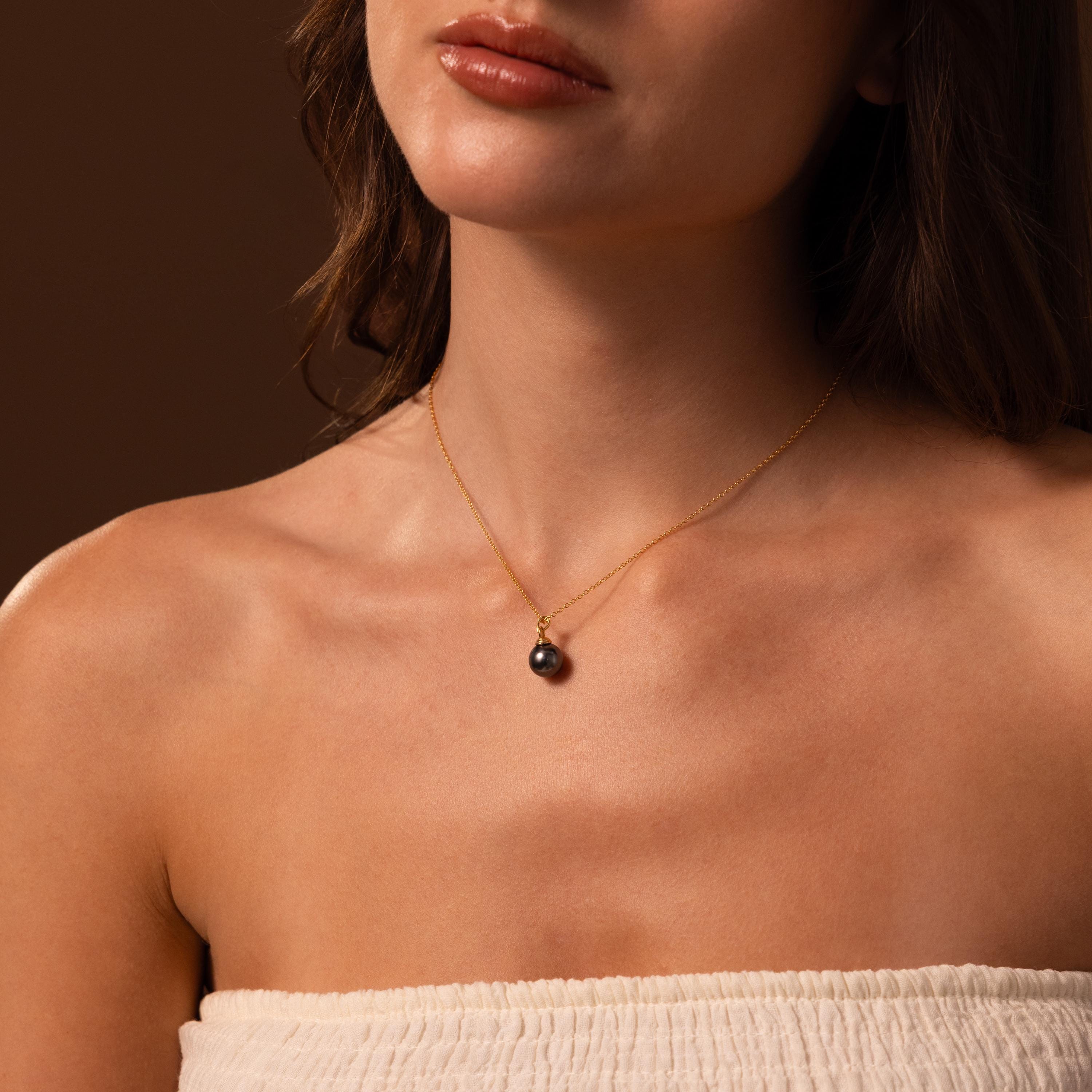 A woman wears the Midnight Pearl Necklace—a gold chain with a black pearl pendant—styled with a white strapless top for a look of timeless sophistication.
