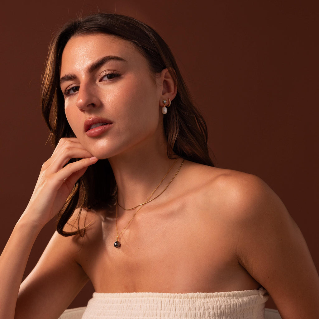 A woman in a strapless top poses against a brown background, wearing Midnight Pearl Drop Earrings and layered necklaces that add timeless elegance to her look.