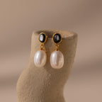The Midnight Pearl Drop Earrings in 18K Gold feature chic gold and black studs with black accents and dangling white pearls, displayed on a textured beige stand—a timeless jewelry piece.
