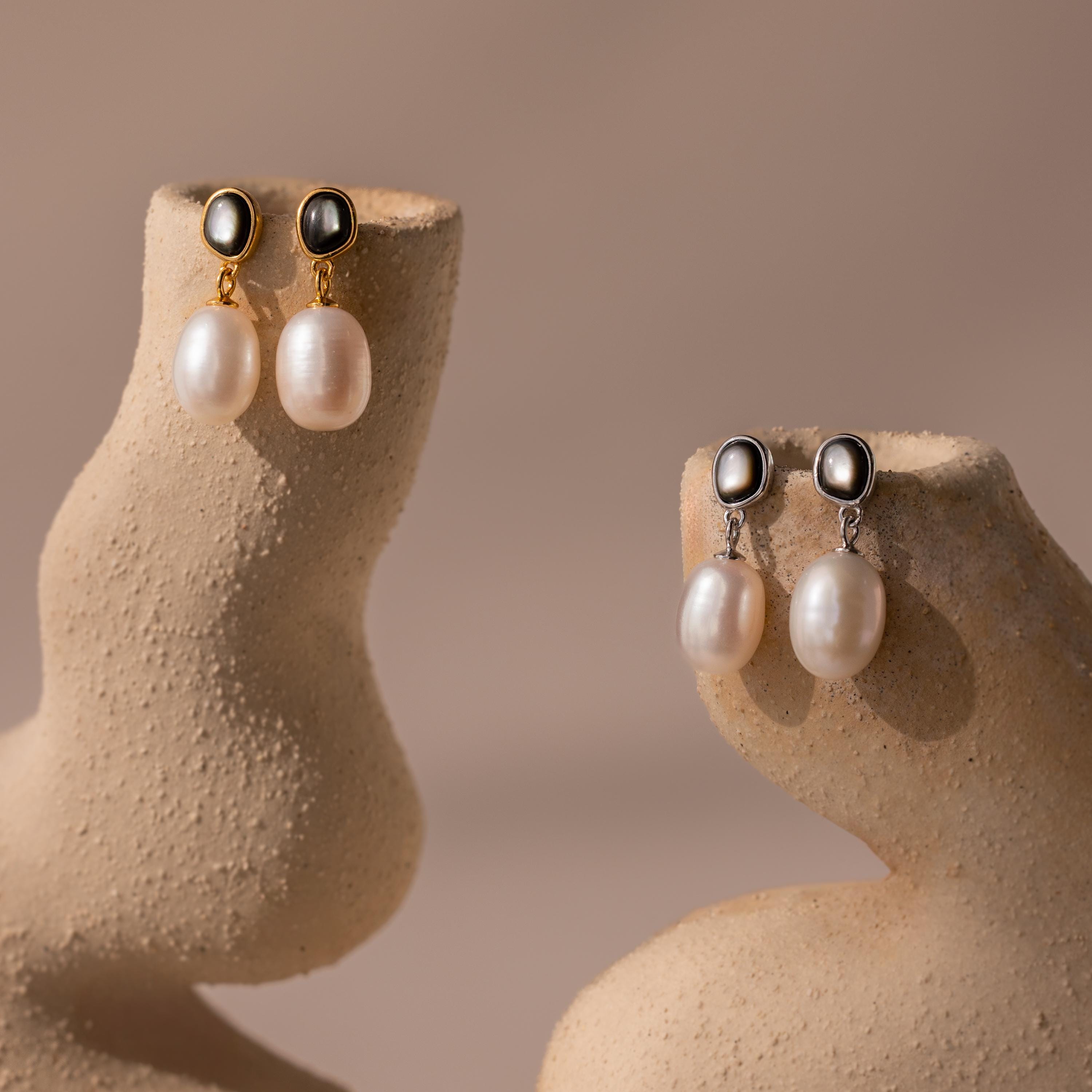 The Midnight Pearl Drop Earrings come in gold and silver styles, each showcasing timeless design with subtle black accents, elegantly presented on textured beige stands.