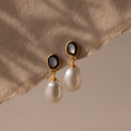 The Midnight Pearl Drop Earrings, featuring gold, pearls, and black accents, rest on a beige textured fabric background and cast soft shadows—a display of timeless jewelry elegance.